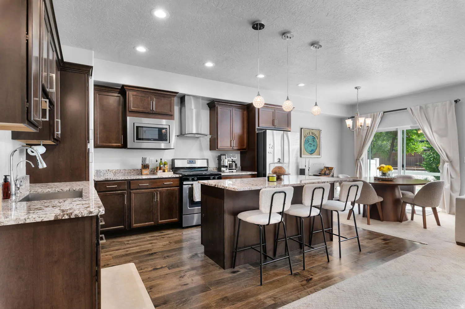 Open-concept kitchen featuring dark wood cabinetry, light granite countertops, stainless steel appliances, a center island with pendant lighting, and wood-finish flooring