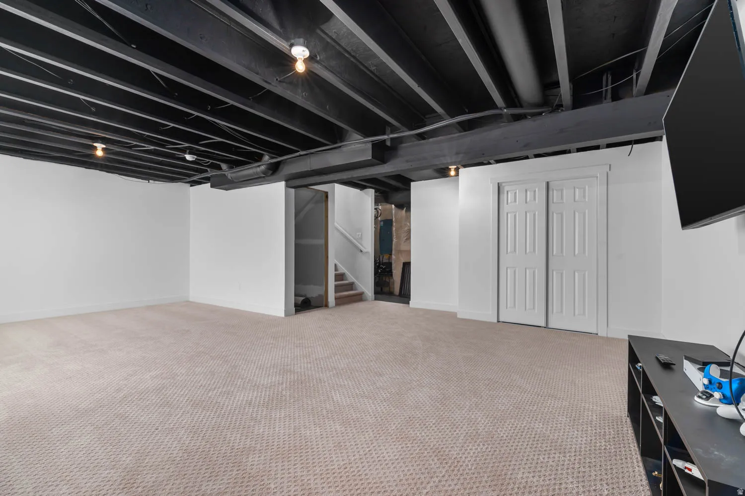 Finished basement space with painted exposed ceiling joists, light-colored carpeting, white painted walls, and built-in closet with paneled bifold doors