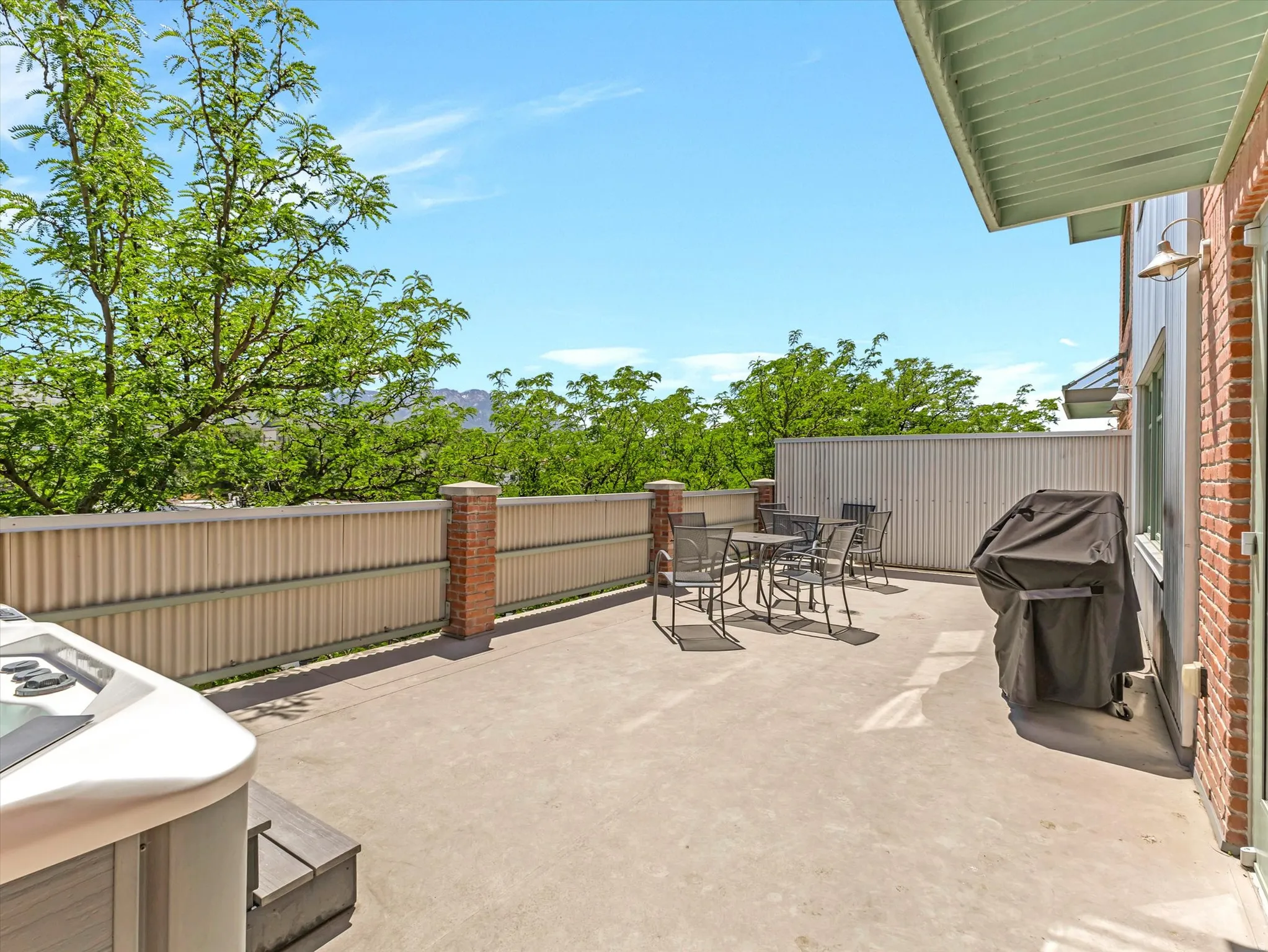 Spacious outdoor patio featuring a hot tub, concrete flooring, corrugated metal fencing with brick pillars, and mature trees