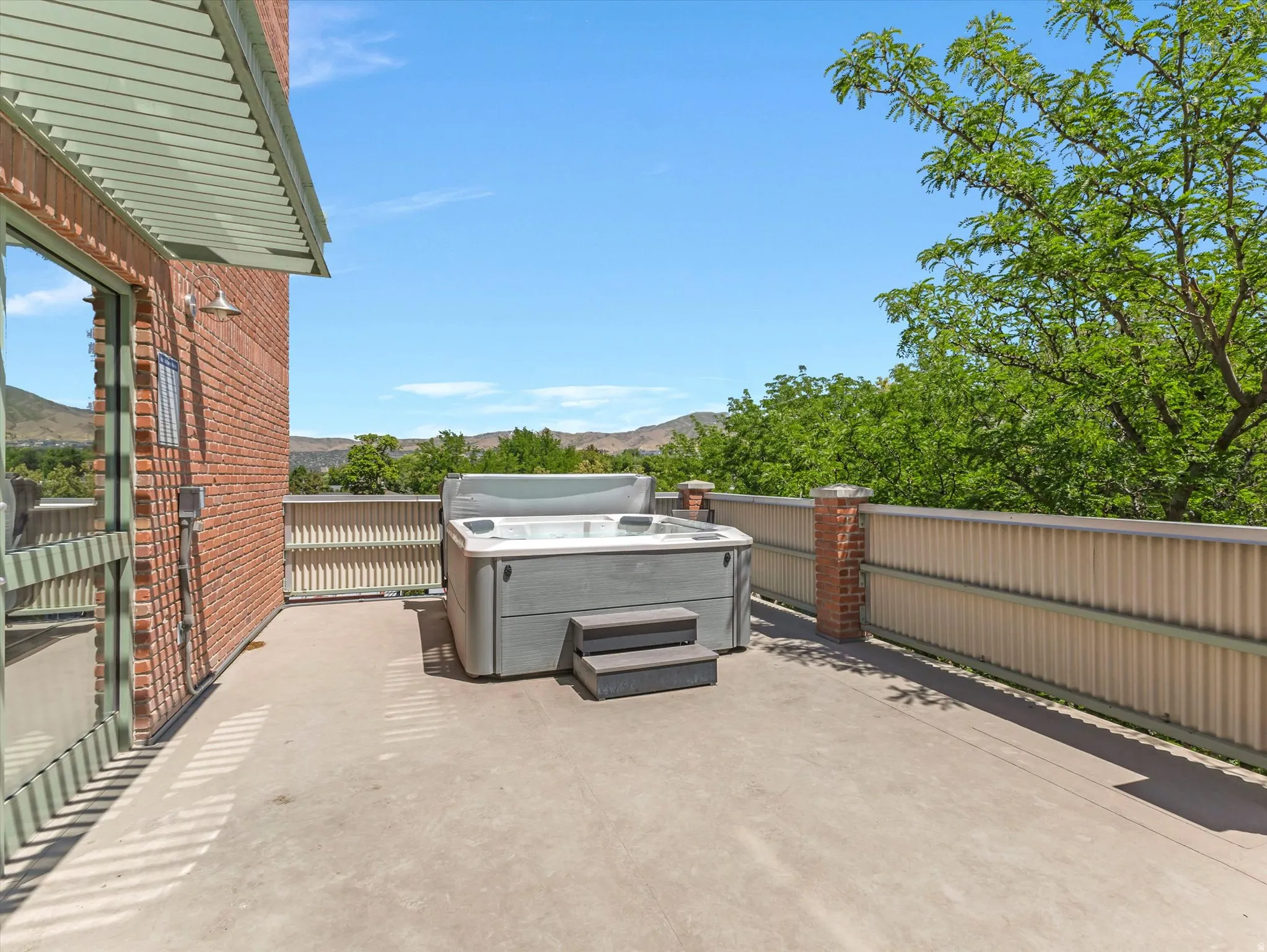 Expansive outdoor patio featuring a hot tub, brick exterior, slatted overhang, and a metal privacy fence