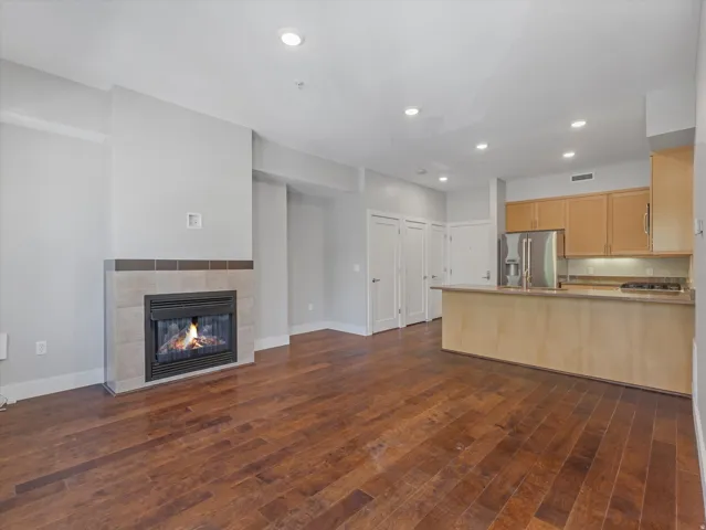 Open-concept living area featuring wood-finish flooring, a tiled fireplace, recessed lighting, and a kitchen with warm-toned cabinetry, stainless steel appliances, and a breakfast bar