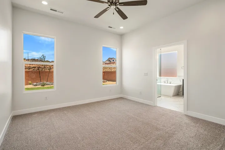Carpeted room featuring two large windows, a ceiling fan, recessed lighting, and a doorway opening to a bathroom with a freestanding tub and glass-enclosed shower