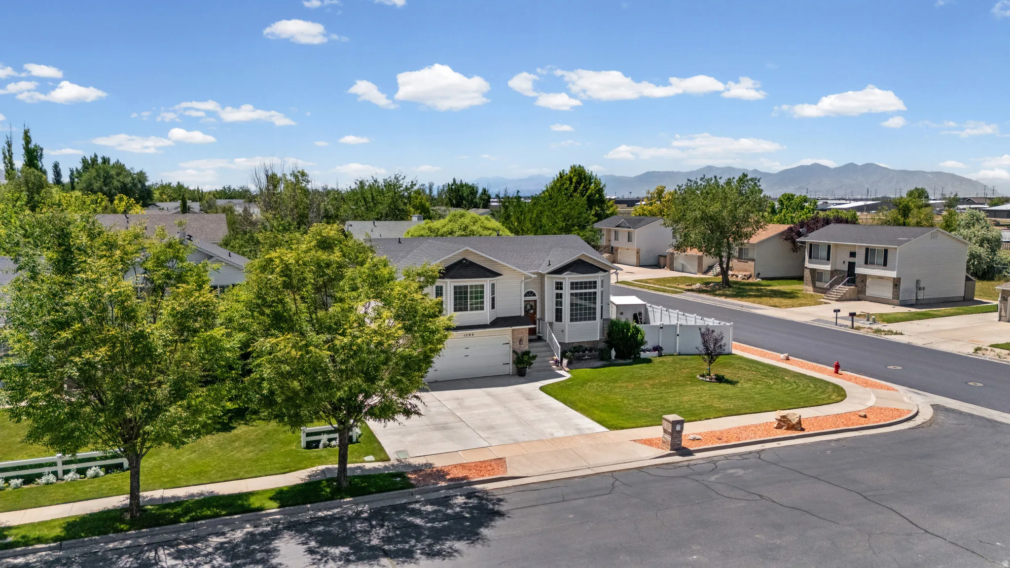 Contemporary residence featuring a two-car garage, concrete driveway, front lawn with mature trees, white picket fence, and a corner lot location