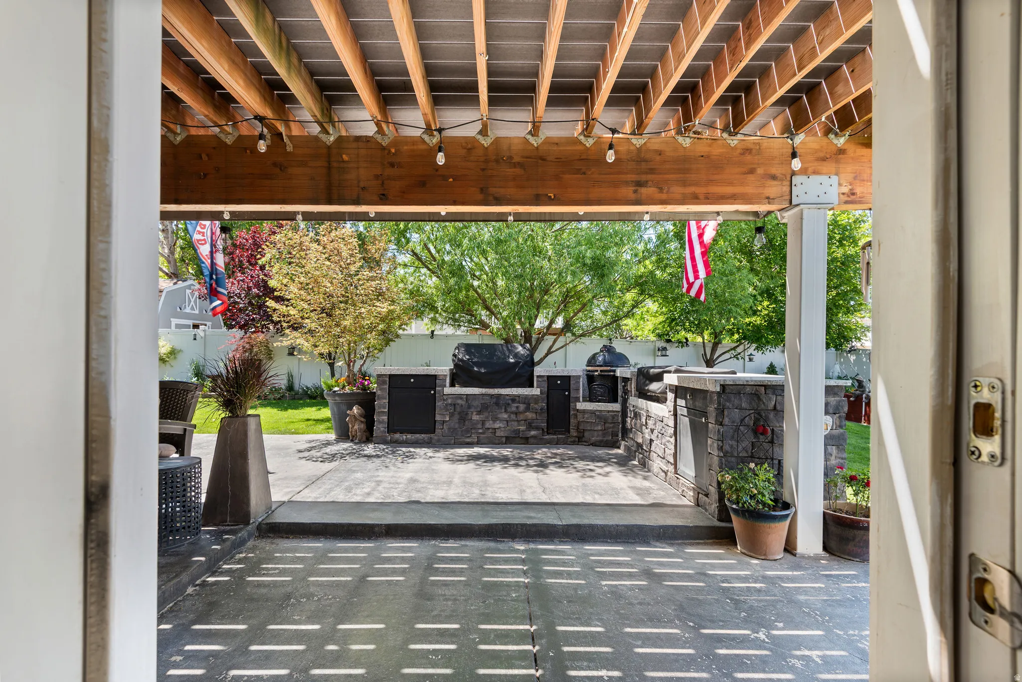 Expansive outdoor patio featuring a built-in stone grill station, covered ceiling with exposed wood beams, string lighting, and a paved surface