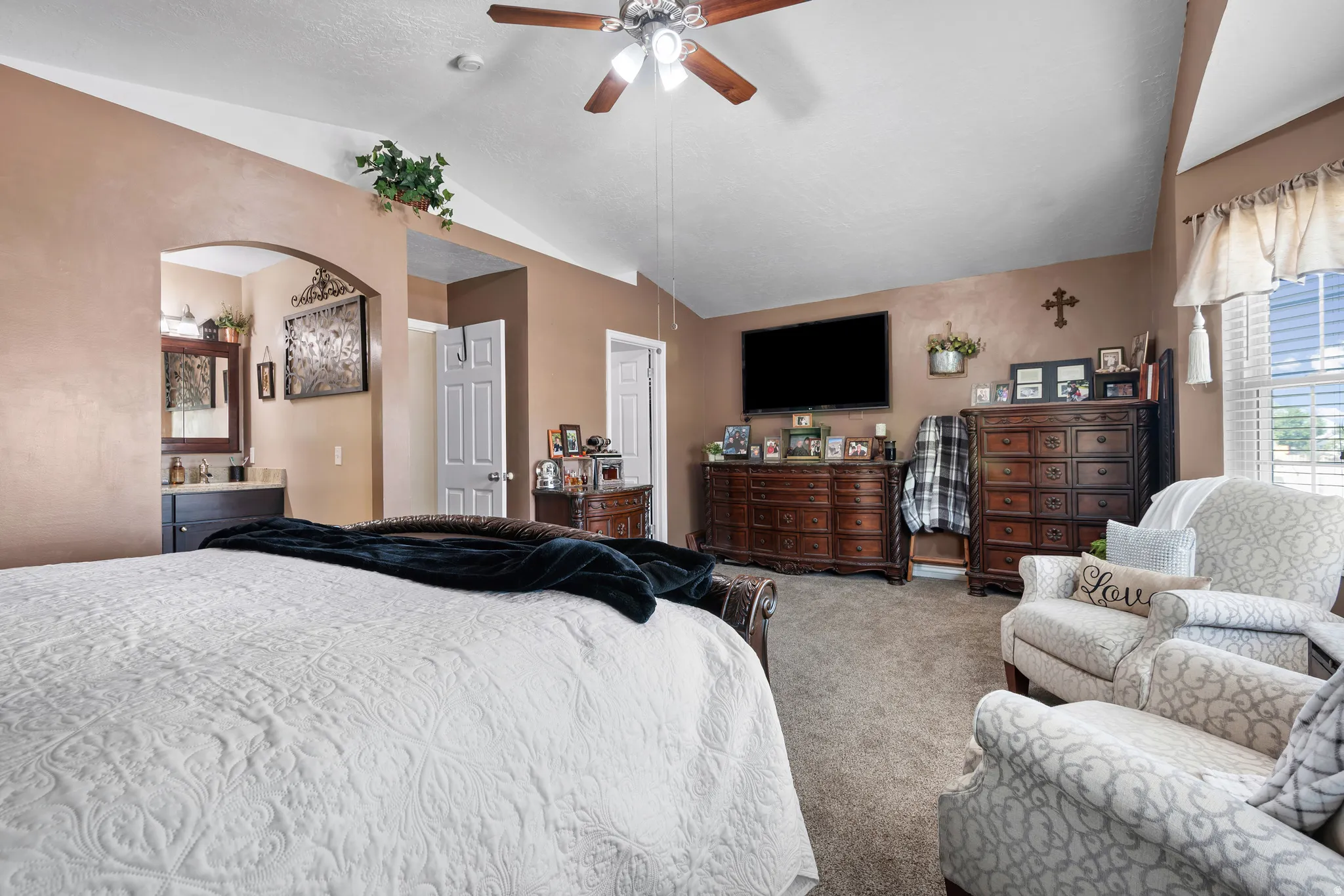 Carpeted room featuring a vaulted ceiling with a ceiling fan, a window with louvered blinds, and an arched doorway leading to a vanity area with a mirror
