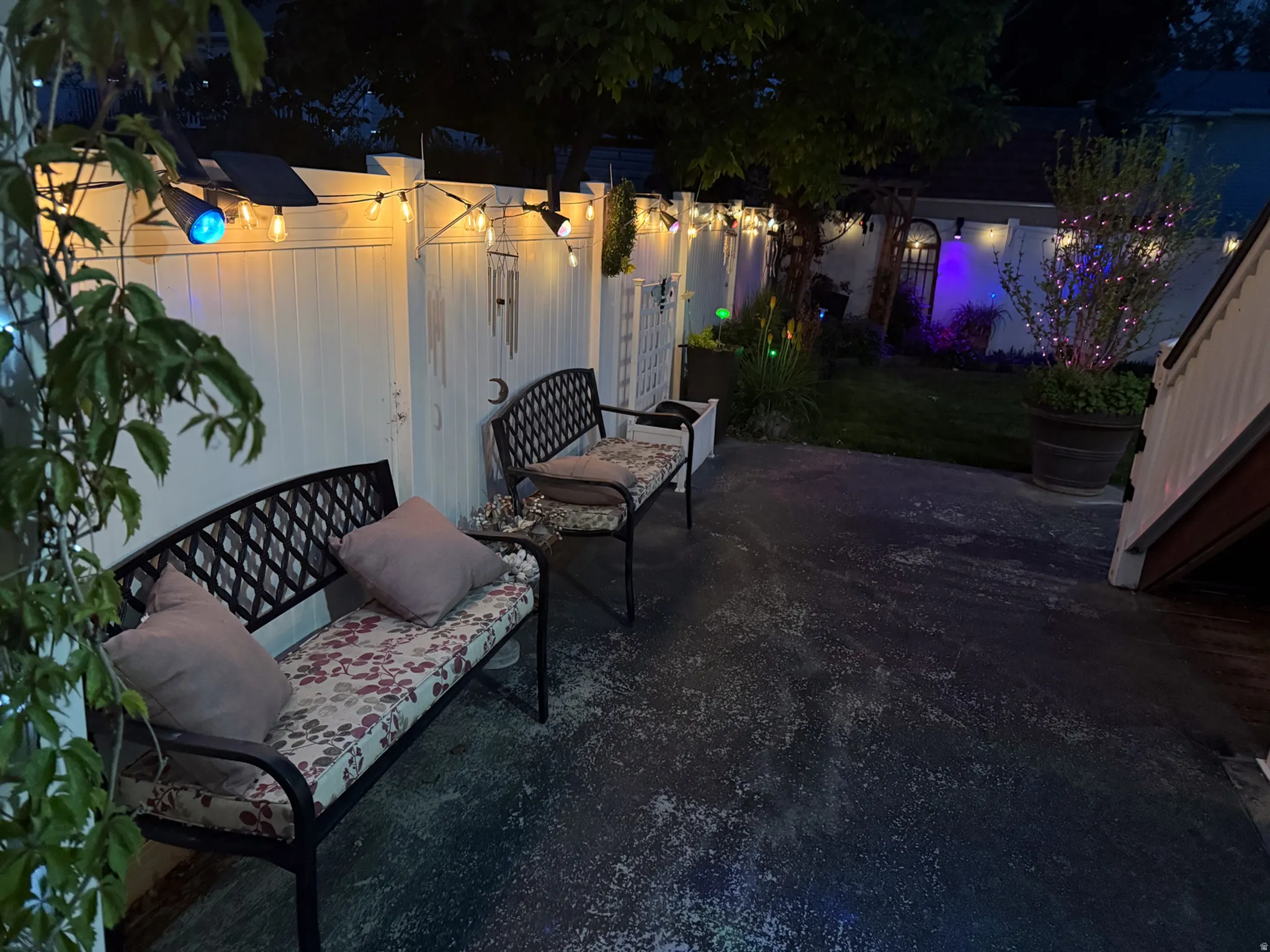 Paved patio space featuring a white privacy fence, string lighting, and adjacent green space