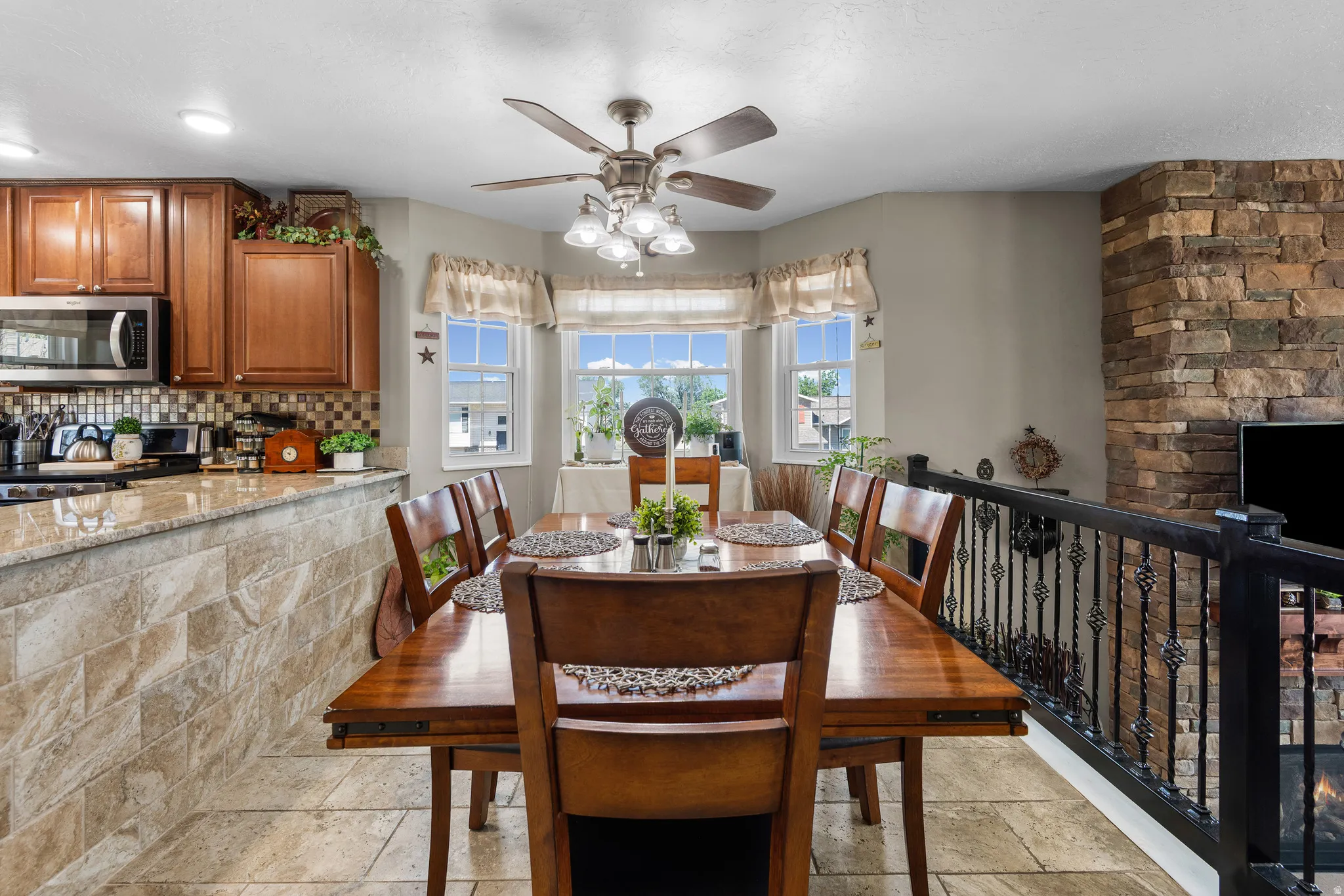 Dining area featuring bay windows, tile flooring, a ceiling fan, a stone-fronted fireplace, and an ornate wrought iron railing