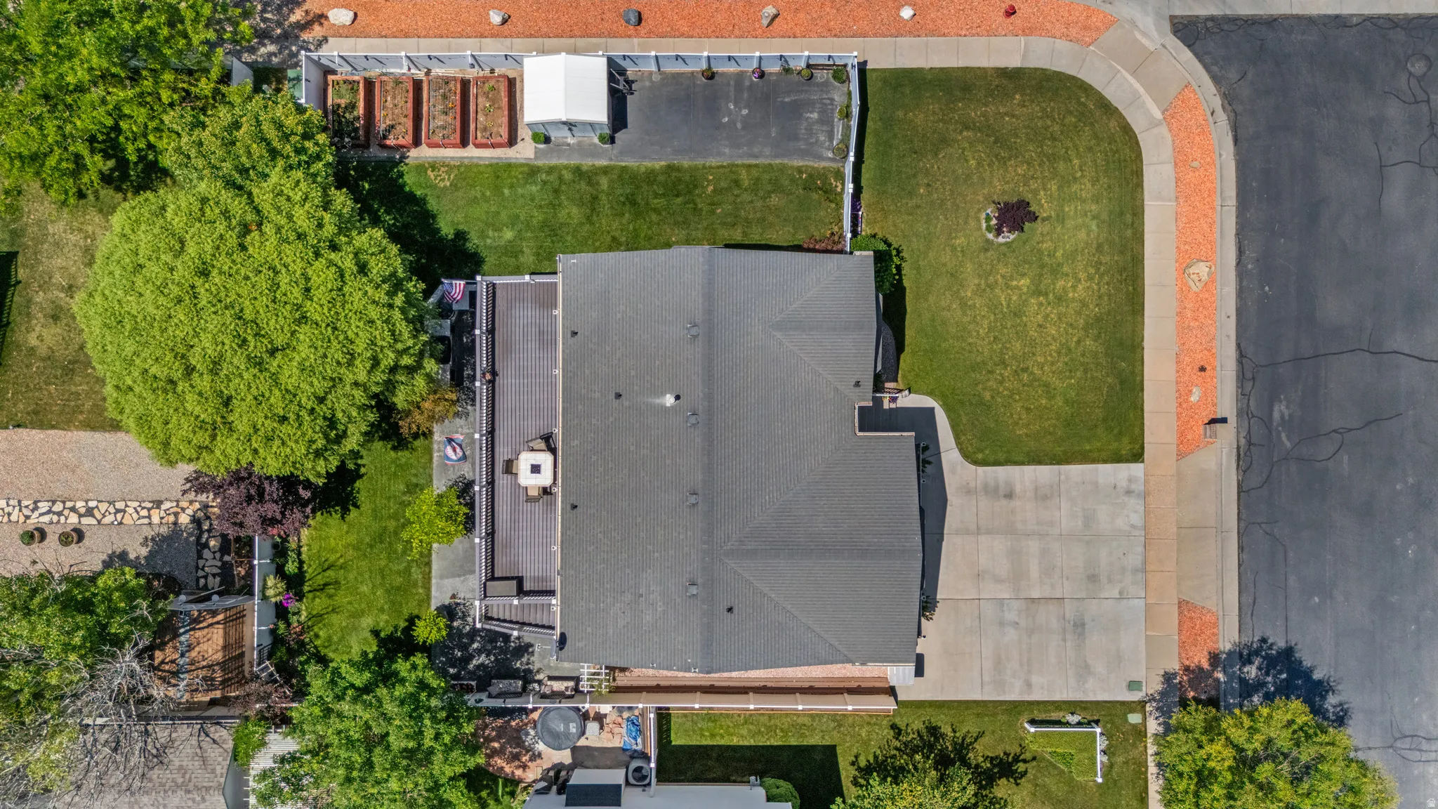Aerial view showcasing a residential property featuring a dark-toned roof, a multi-level deck, a concrete driveway, and a landscaped front yard with a curved lawn