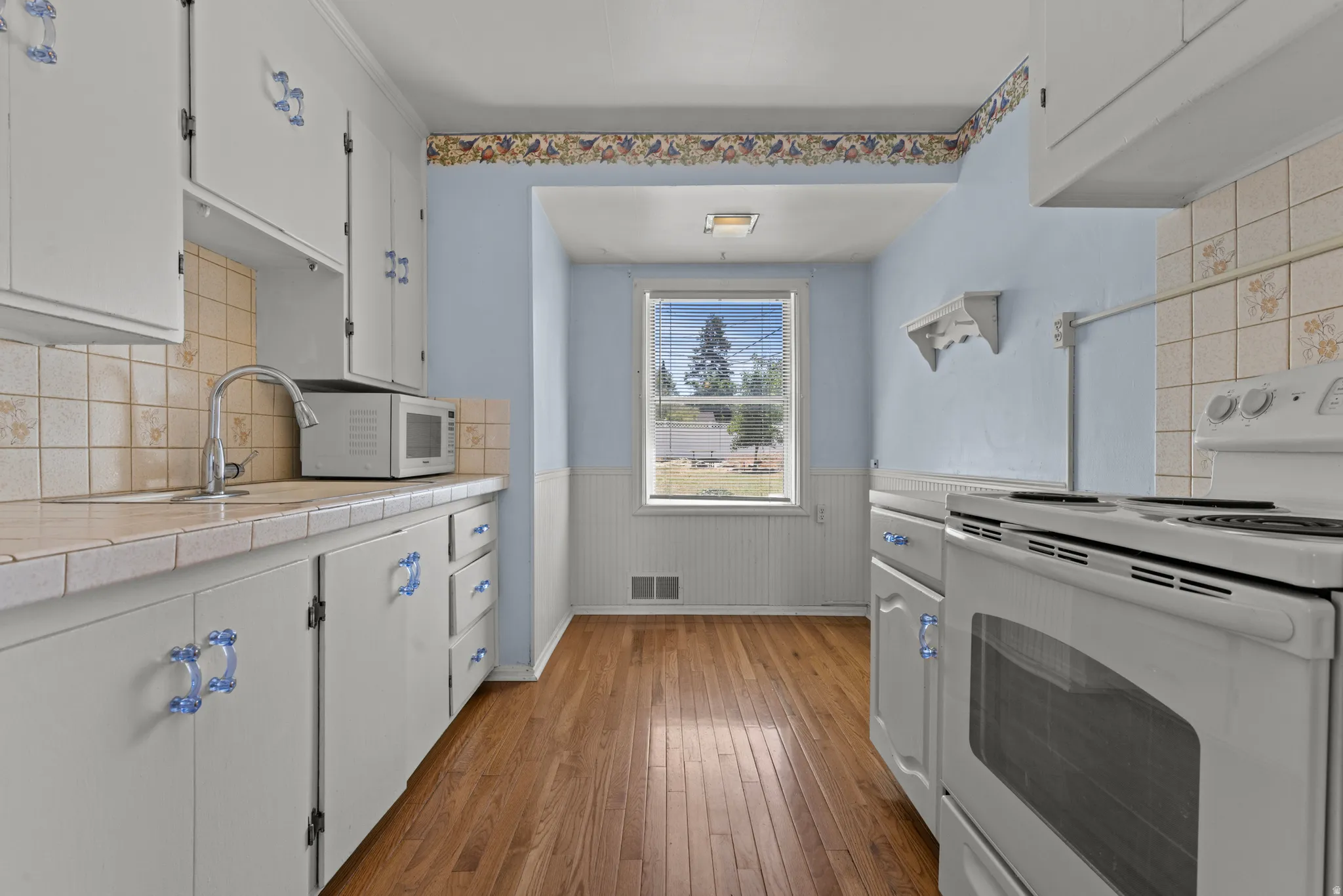 Kitchen featuring wood-finish flooring, white cabinetry with blue decorative pulls, a white electric range, and a window with exterior views