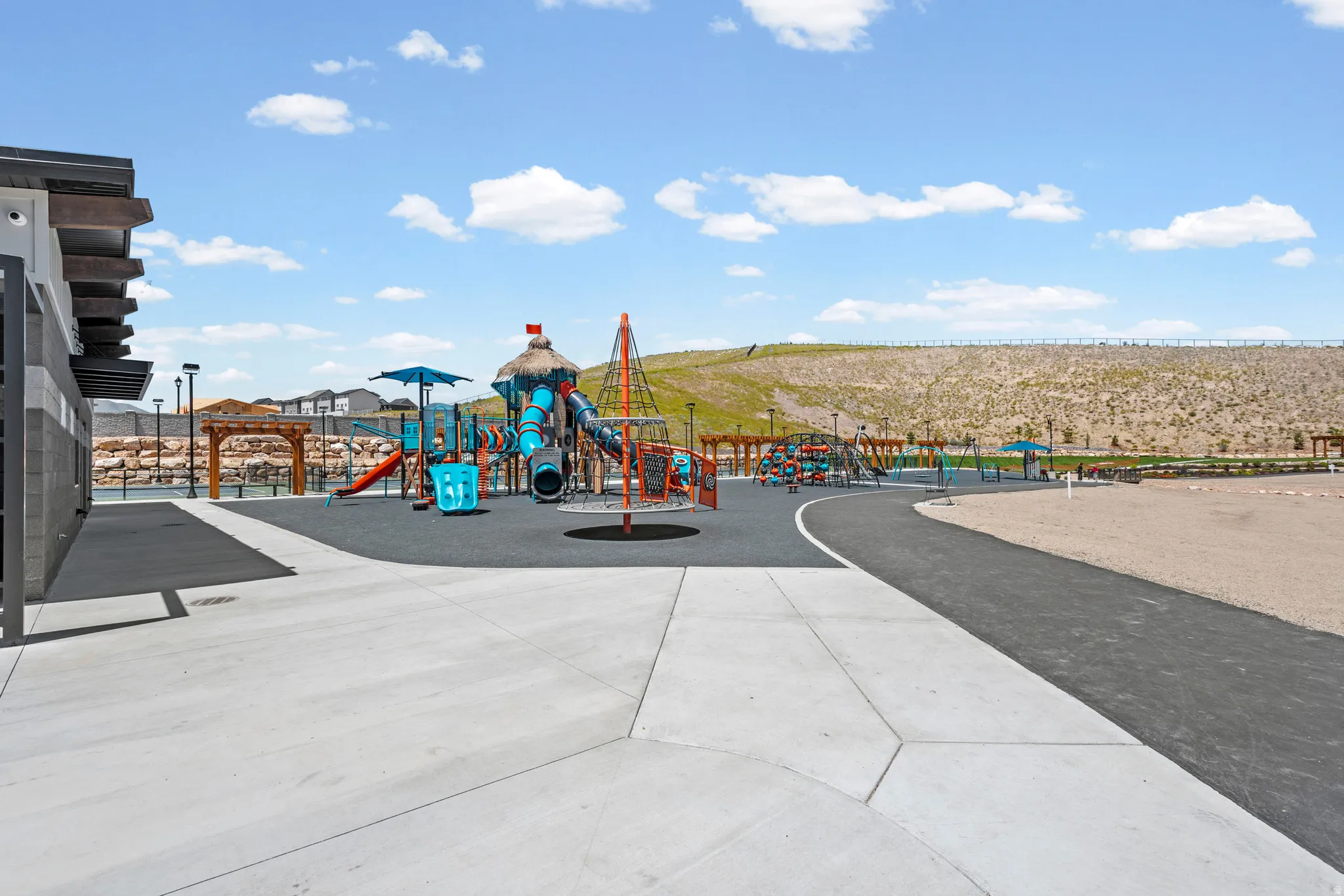 Expansive outdoor recreation area featuring a large playground with multiple slides and climbing structures, a dedicated rubberized surfacing, a paved walking path, a sand play area, and an adjacent pergola structure