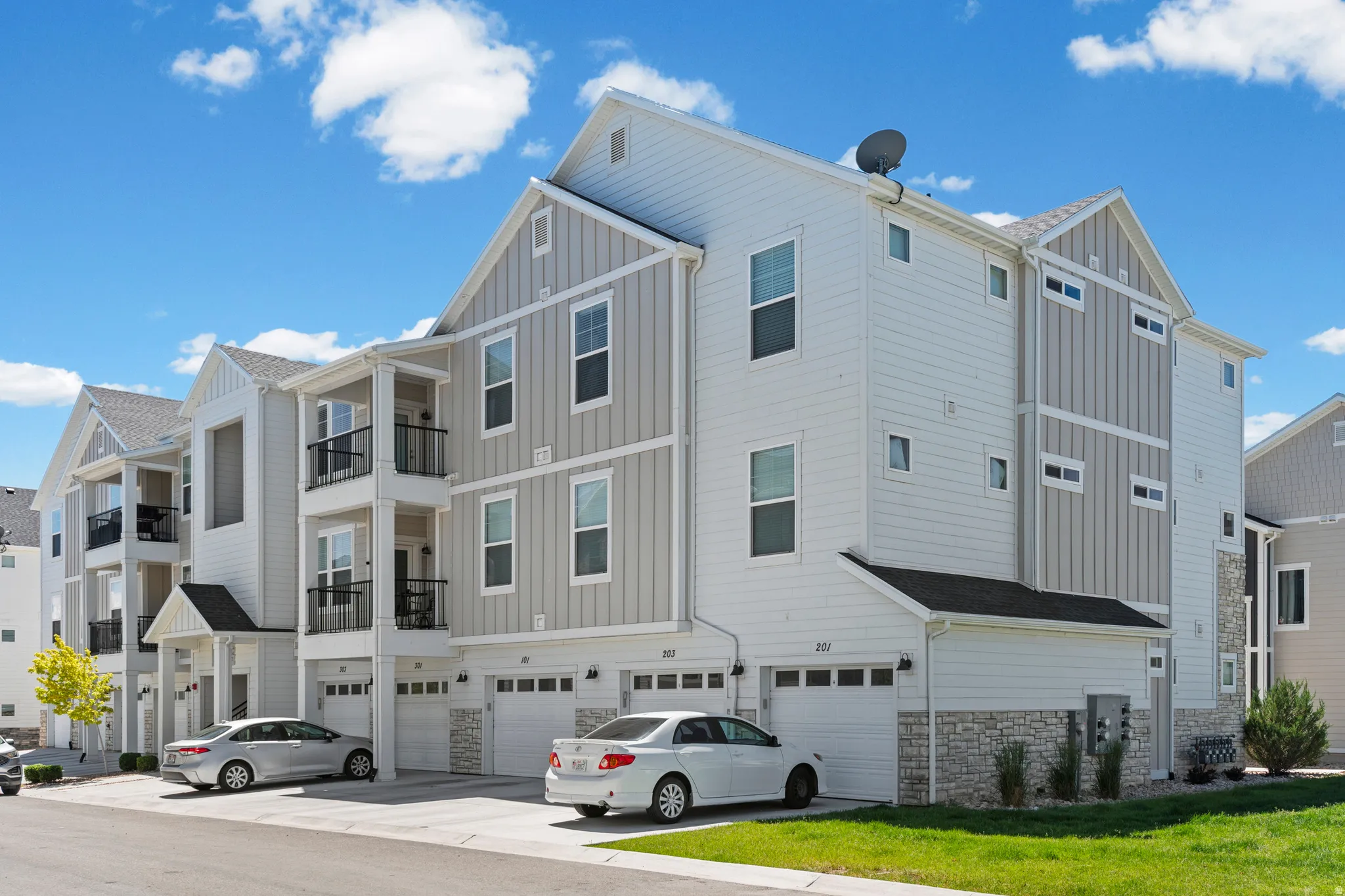 Modern townhome exterior featuring light-colored siding, a stone facade accent, individual garages, and private balconies with black railings
