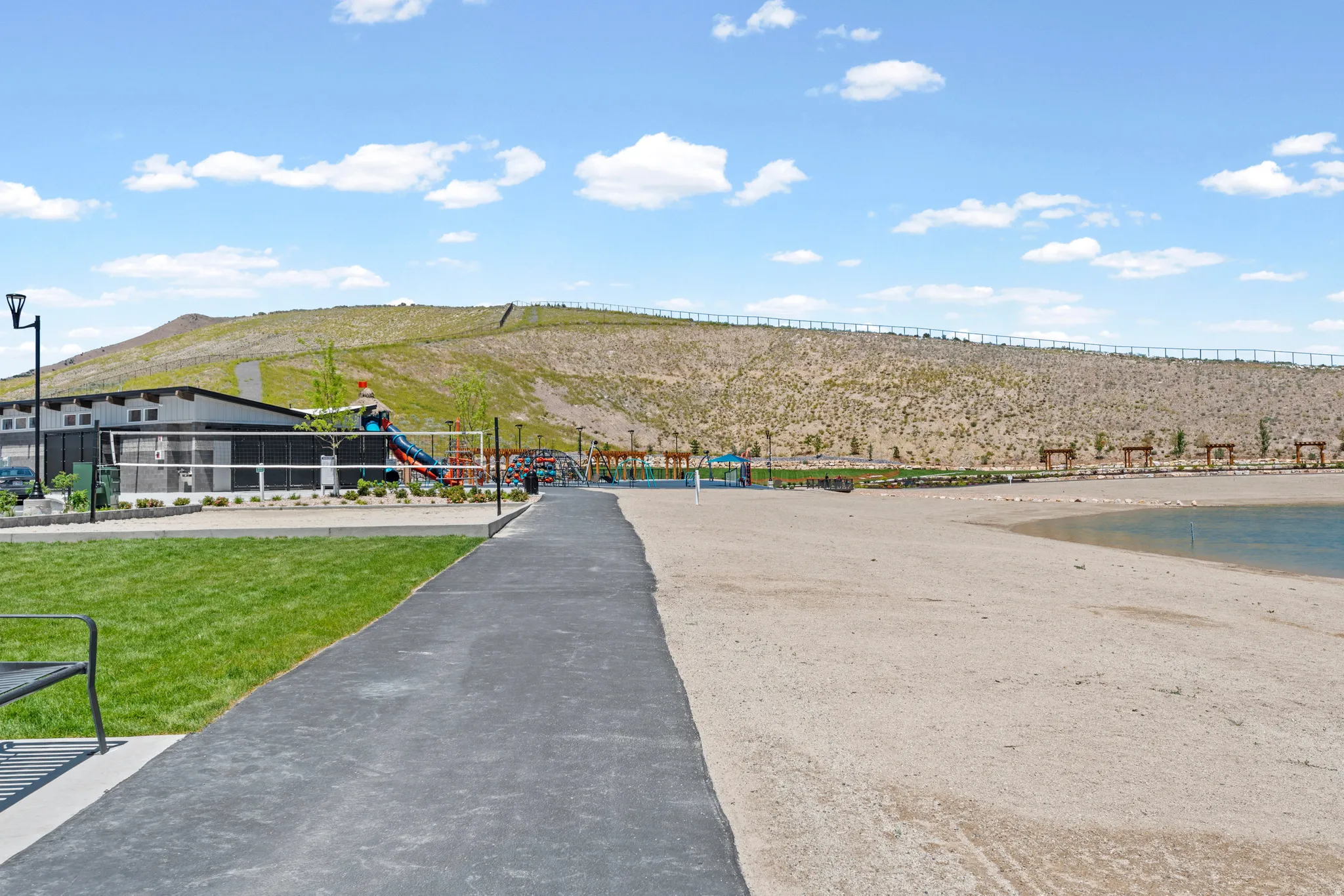 Recreational area featuring a sandy beach, paved pathway, green lawn, a contemporary building, and playground equipment