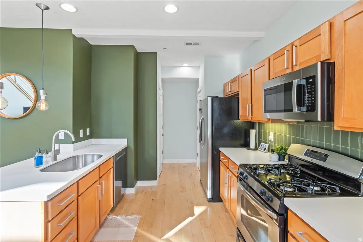 Modern kitchen featuring light wood-finish cabinetry, stainless steel appliances, white countertops, and a green tiled backsplash