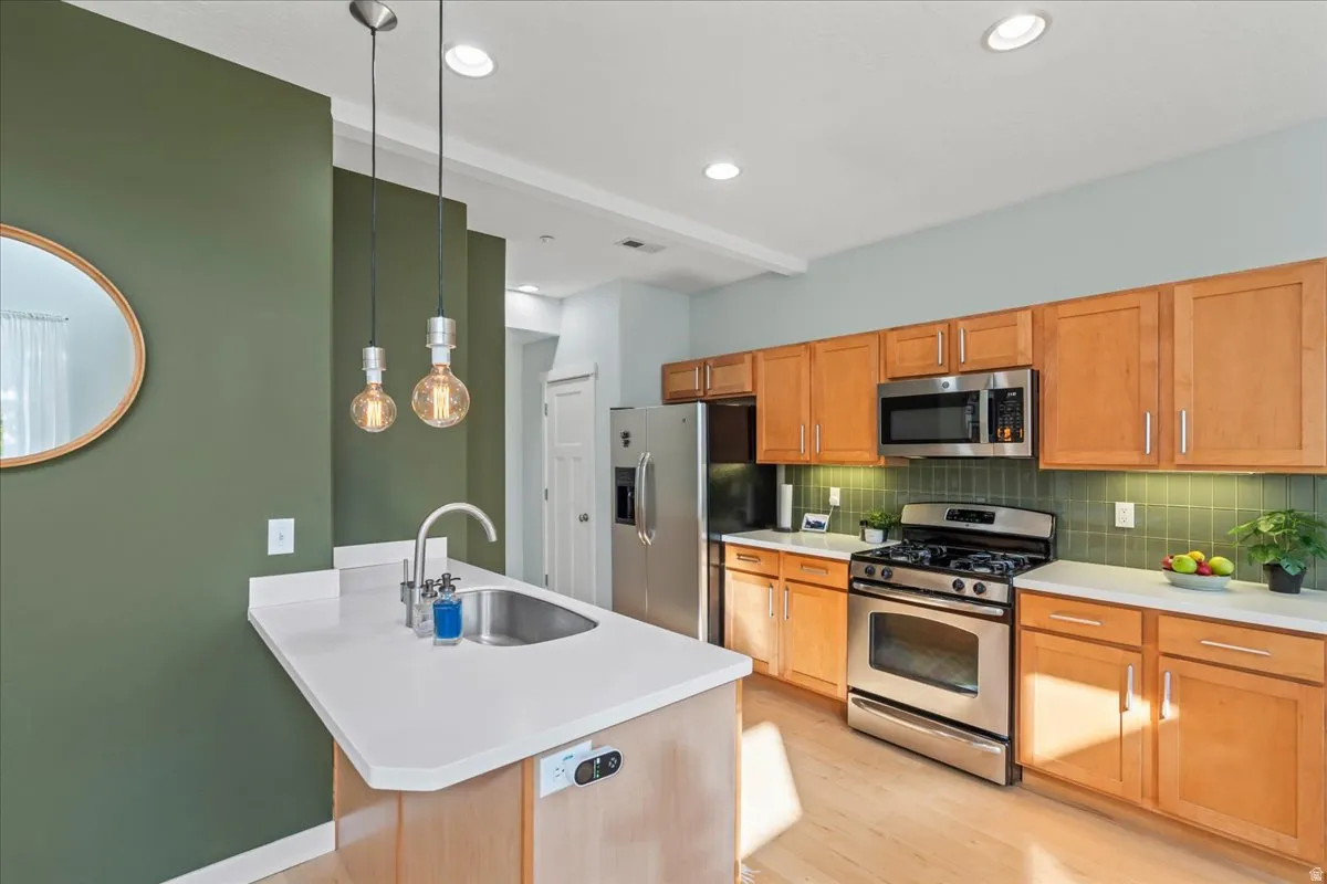 Modern kitchen featuring a central island with an undermount sink and pendant lighting, light wood-finish flooring, stainless steel appliances, light wood cabinetry, and a subway tile backsplash