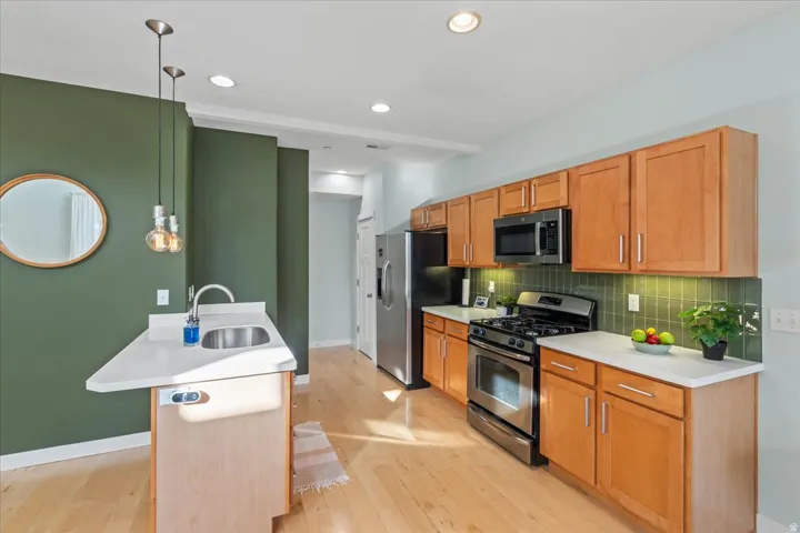 Kitchen featuring light wood-finish flooring, light wood cabinetry, stainless steel appliances, white countertops, and a green tile backsplash