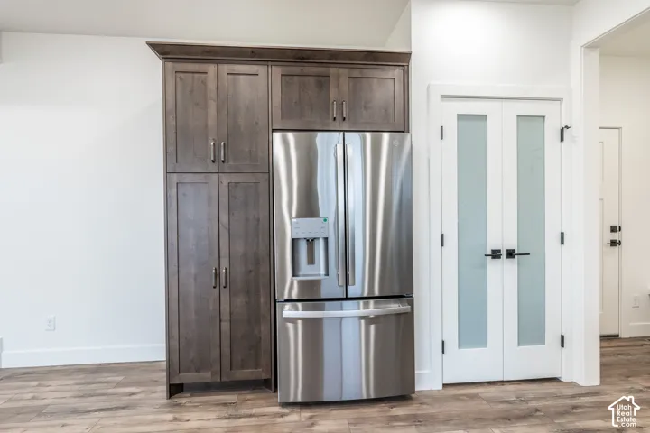 Stainless steel French door refrigerator, tall wood-finish cabinetry, white shaker-style double doors with frosted glass, matte black hardware, and wood-finish flooring