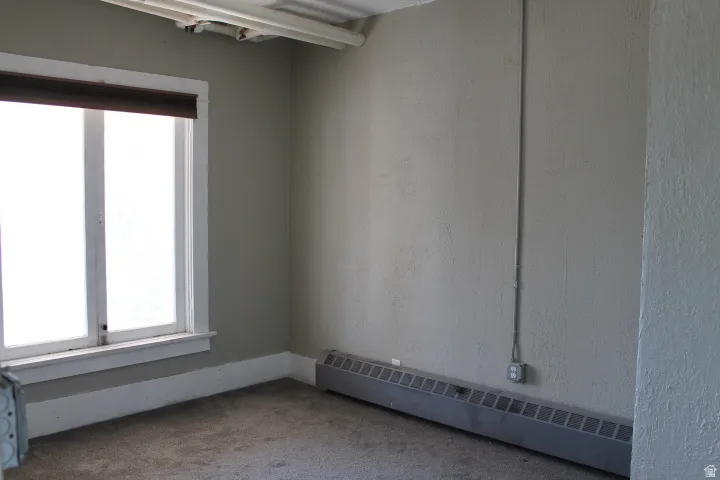 Interior space featuring a large window with white trim, a dark roller shade, baseboard heating unit, and light gray walls