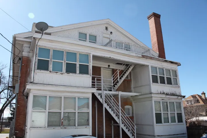 Multi-story building featuring brick and white-painted siding, multiple enclosed sunrooms, exterior staircases, and a prominent brick chimney