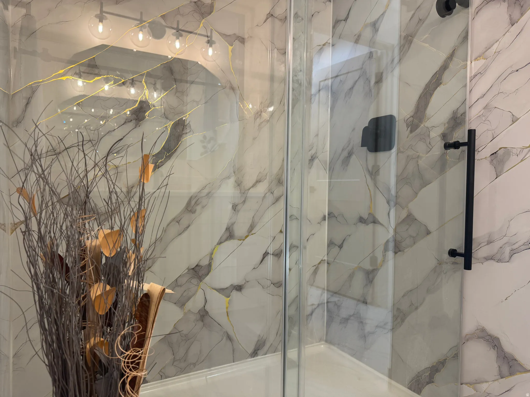 Spacious glass-enclosed shower with white and grey marbled wall tiling, black hardware, and a rainfall shower head