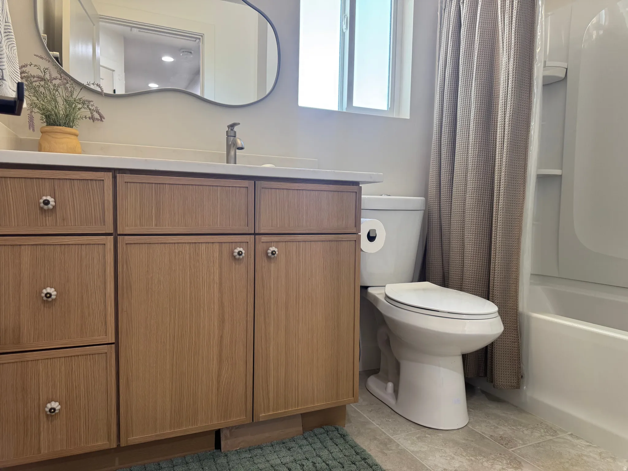 Bathroom featuring a wood-finish vanity with drawer and cabinet storage, a white countertop, a contemporary mirror, a full bathtub with shower, and light-toned tile flooring