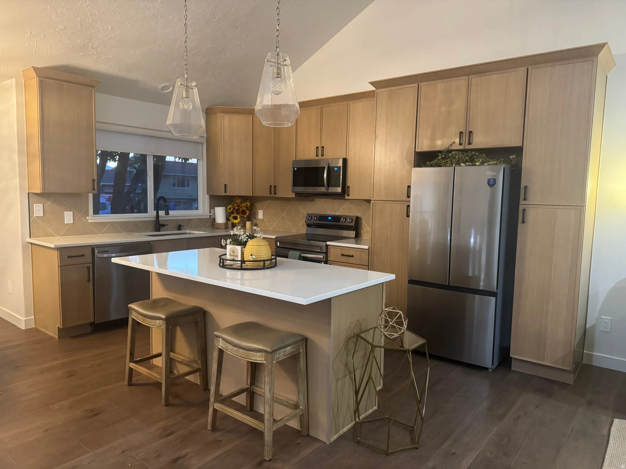Kitchen featuring an island with white countertop, stainless steel appliances, wood-finish flooring, light wood cabinetry, and a tile backsplash