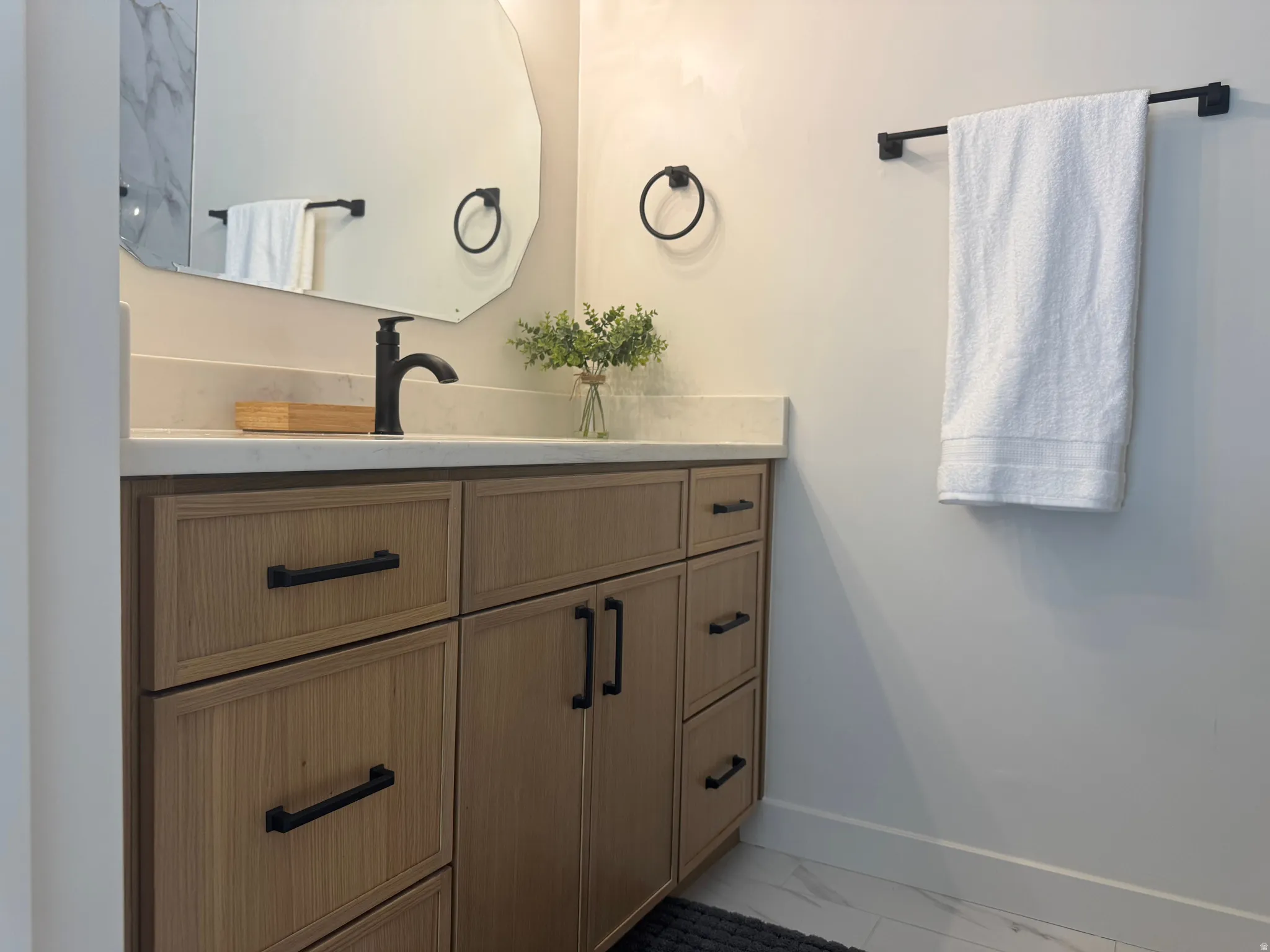 Bathroom vanity featuring light wood-finish cabinetry with black hardware, a light-toned countertop, a black single-handle faucet, and an irregular-shaped mirror