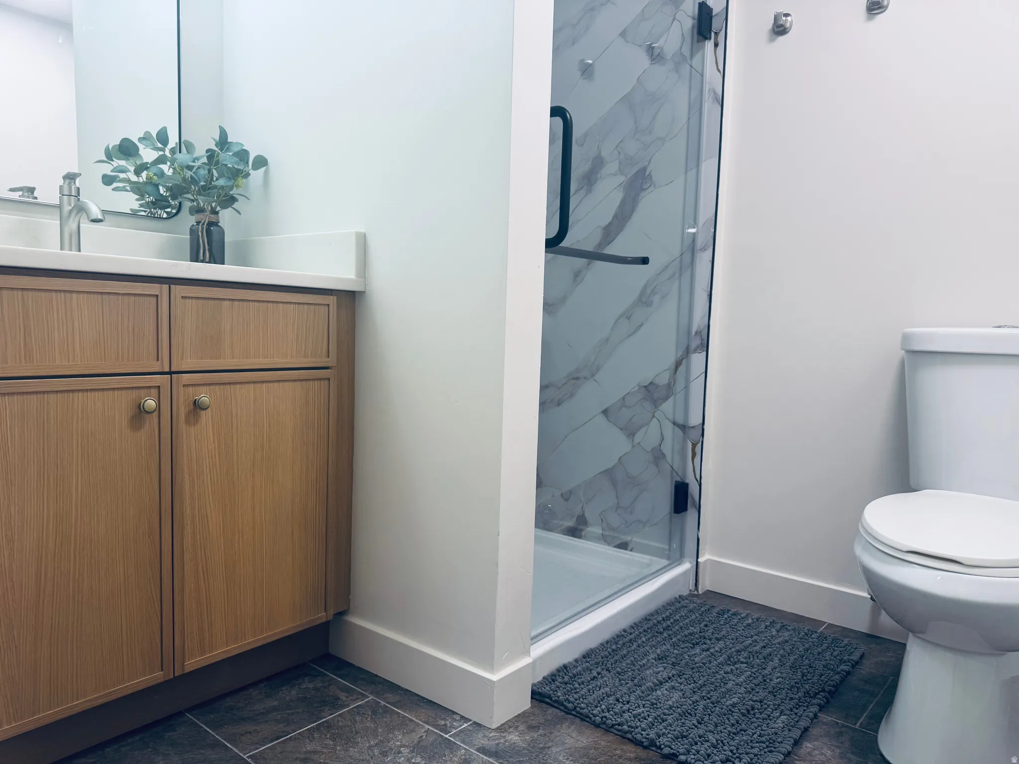 Bathroom featuring a wood-finish vanity with a light-colored countertop, a chrome faucet, a glass-enclosed shower with large format marble-look tile, and a white toilet