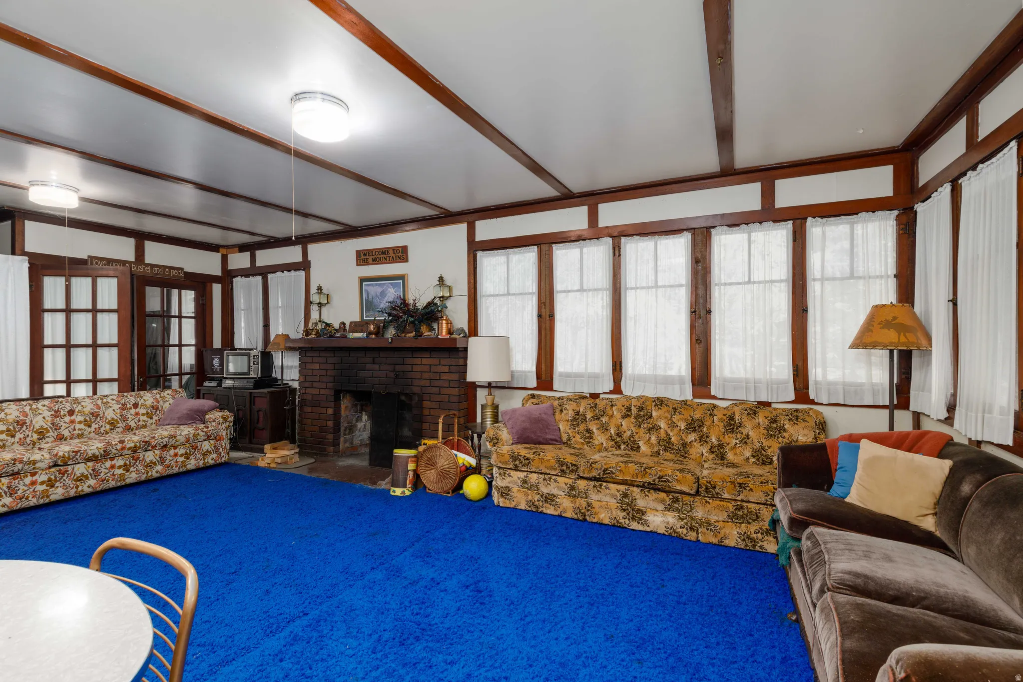 Spacious interior room featuring a prominent brick fireplace with a mantel, multiple windows, double French doors, and exposed ceiling beams