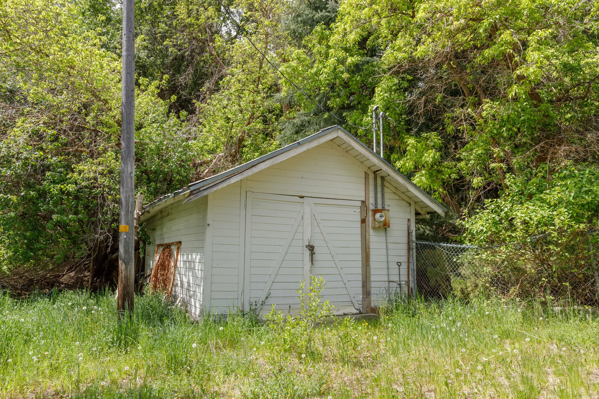 White horizontal siding structure with a pitched roof and double front doors, surrounded by mature trees and overgrown grass