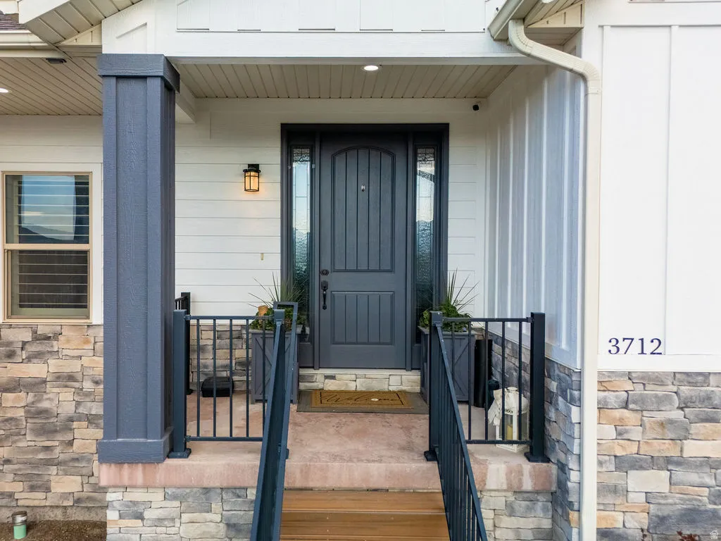 Inviting front entry featuring a dark paneled door with glass sidelights, board and batten siding, stone veneer accents, and a covered porch