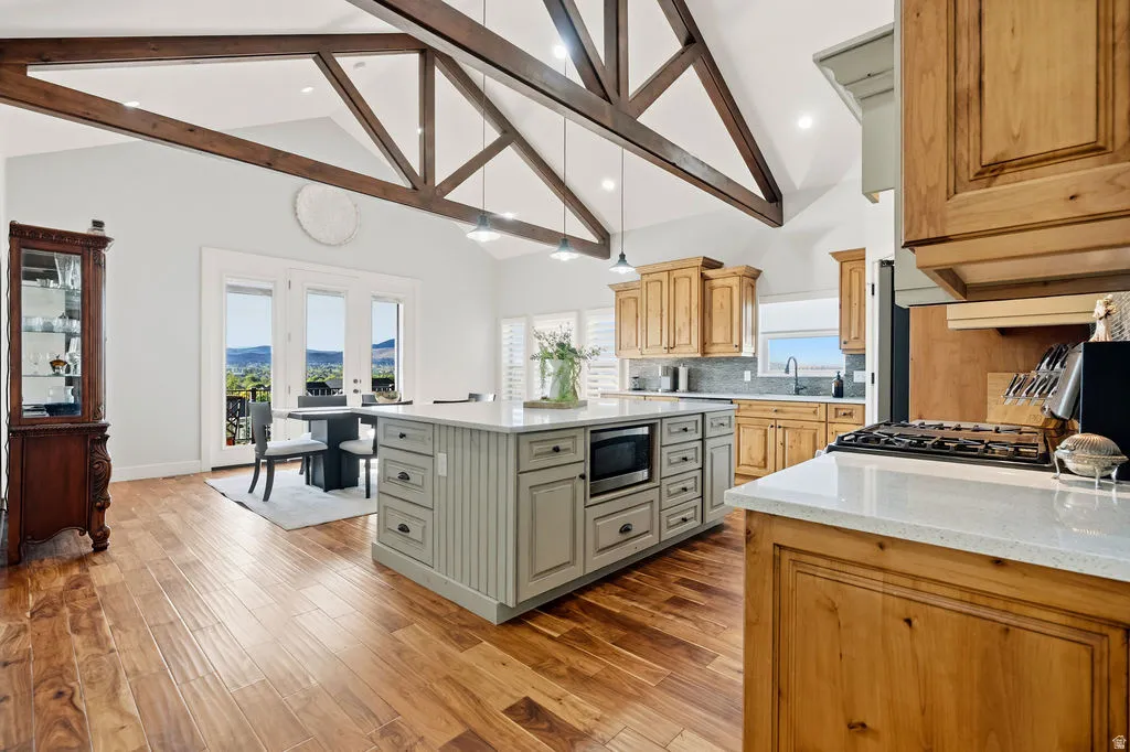 Spacious kitchen featuring a large central island with a built-in microwave, wood-finish flooring, exposed beam vaulted ceilings, light wood cabinetry, and a gas cooktop