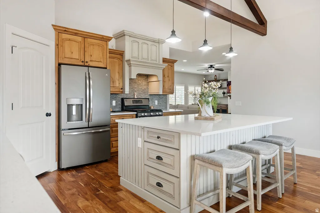Spacious kitchen featuring a large island with a light-toned countertop, stainless steel appliances, wood-finish flooring, exposed ceiling beams, and pendant lighting