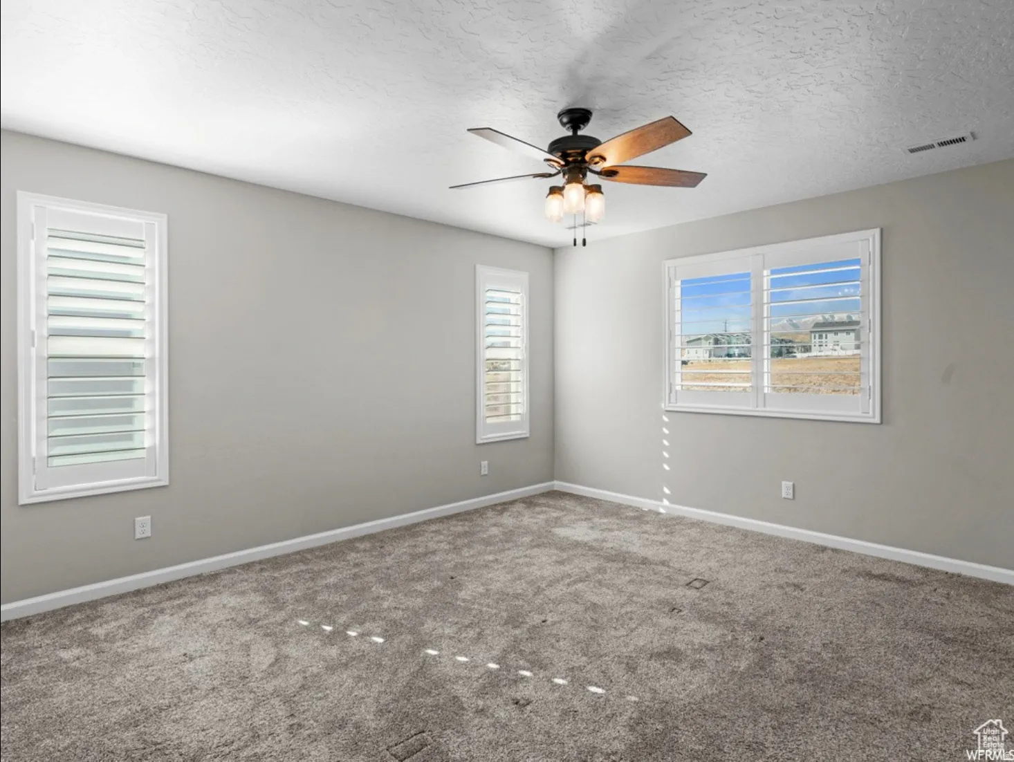 Spacious room featuring neutral paint, gray carpet, three windows with white plantation shutters, and a ceiling fan with integrated lighting