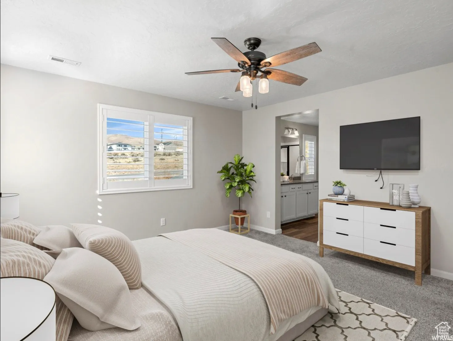 Carpeted bedroom featuring a ceiling fan with light fixture, window with plantation shutters, and an entryway to a bathroom with a double vanity