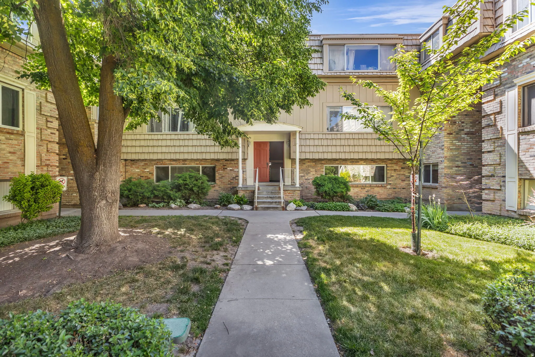 Building exterior featuring brick and shingle siding, a red front door, concrete walkway, and established landscaping