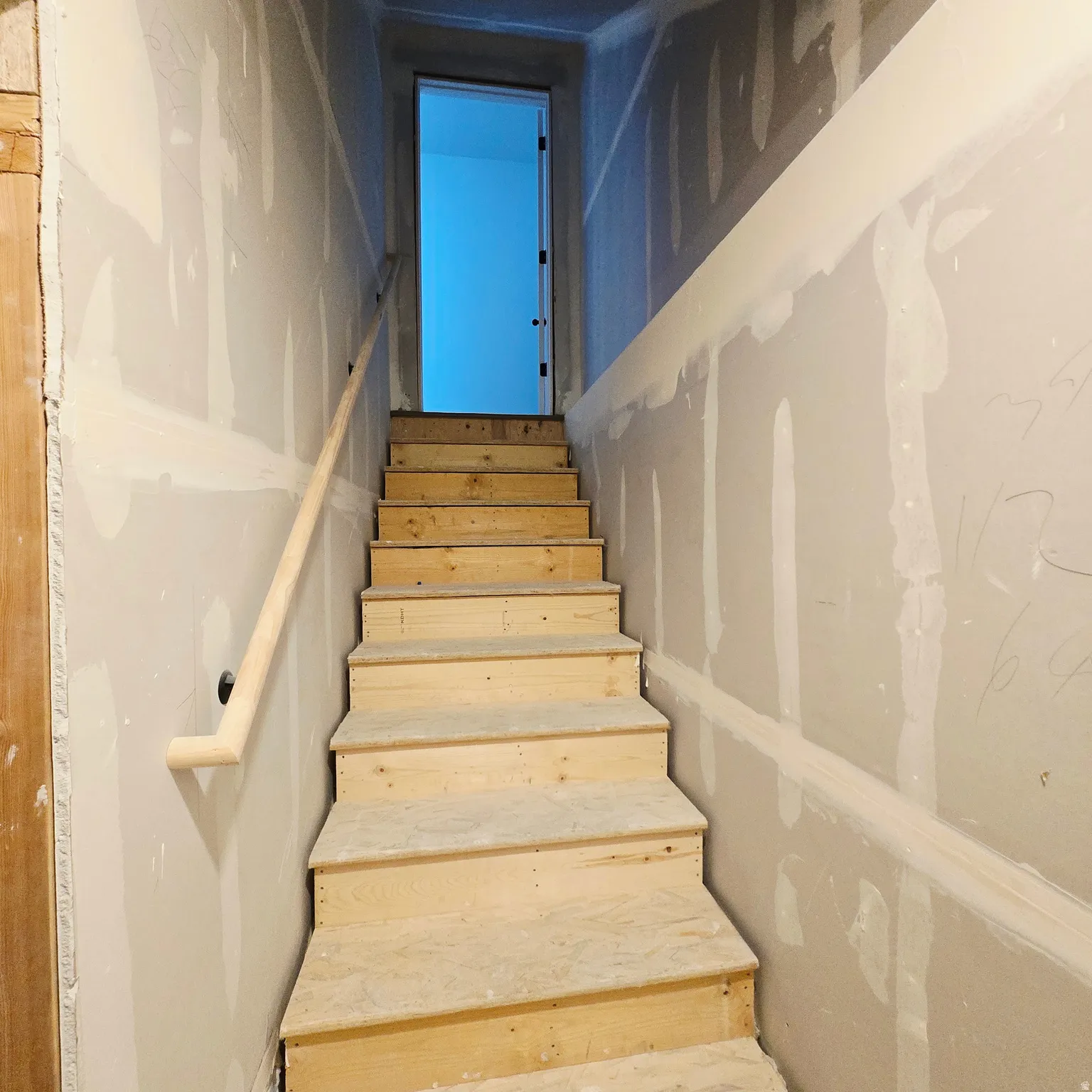 Unfinished wooden staircase with a simple wood handrail, drywall-finished walls, and an open doorway at the top