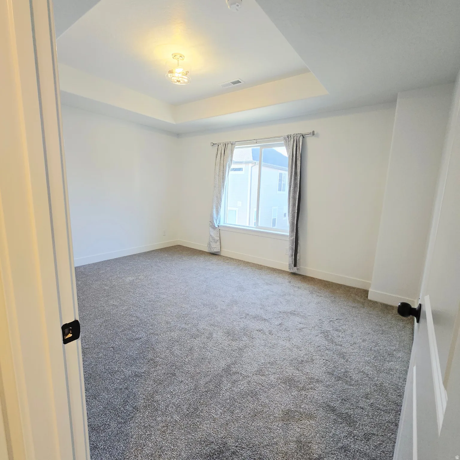 Spacious room featuring a tray ceiling with a central light fixture, a large window with drapes, and plush grey carpeting
