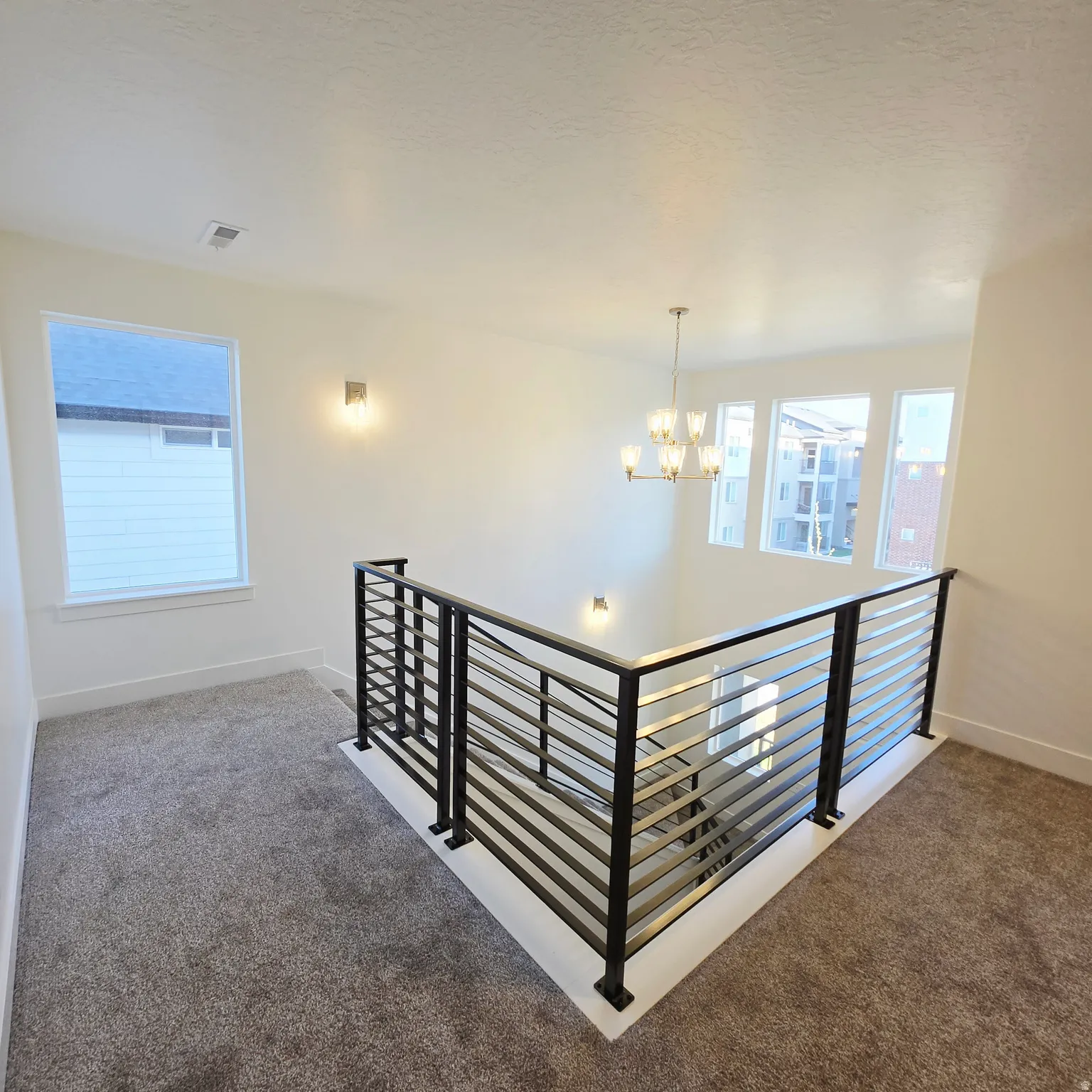 Upper landing featuring contemporary horizontal metal railings, gray carpet, a multi-pane window, and a modern gold-finish chandelier