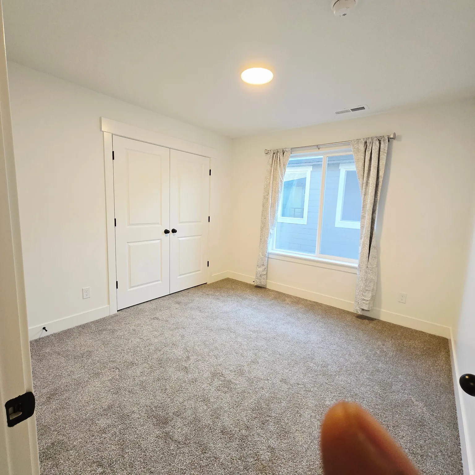 Light-filled room featuring neutral-toned carpeting, white trim, a double-door closet with black hardware, and a window with patterned drapes