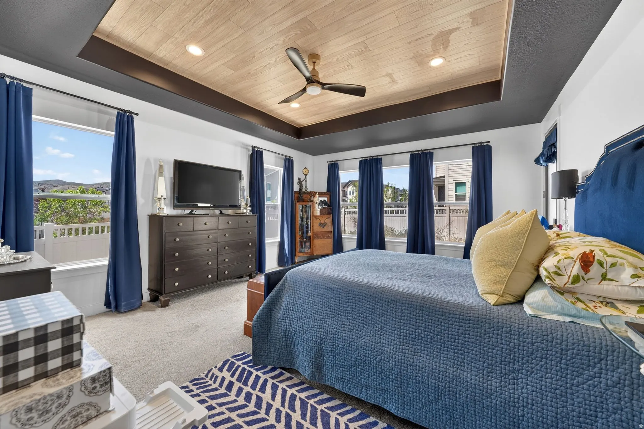 Room featuring a tray ceiling with wood-finish inlay and recessed lighting, multiple windows, soft carpet flooring, and a ceiling fan