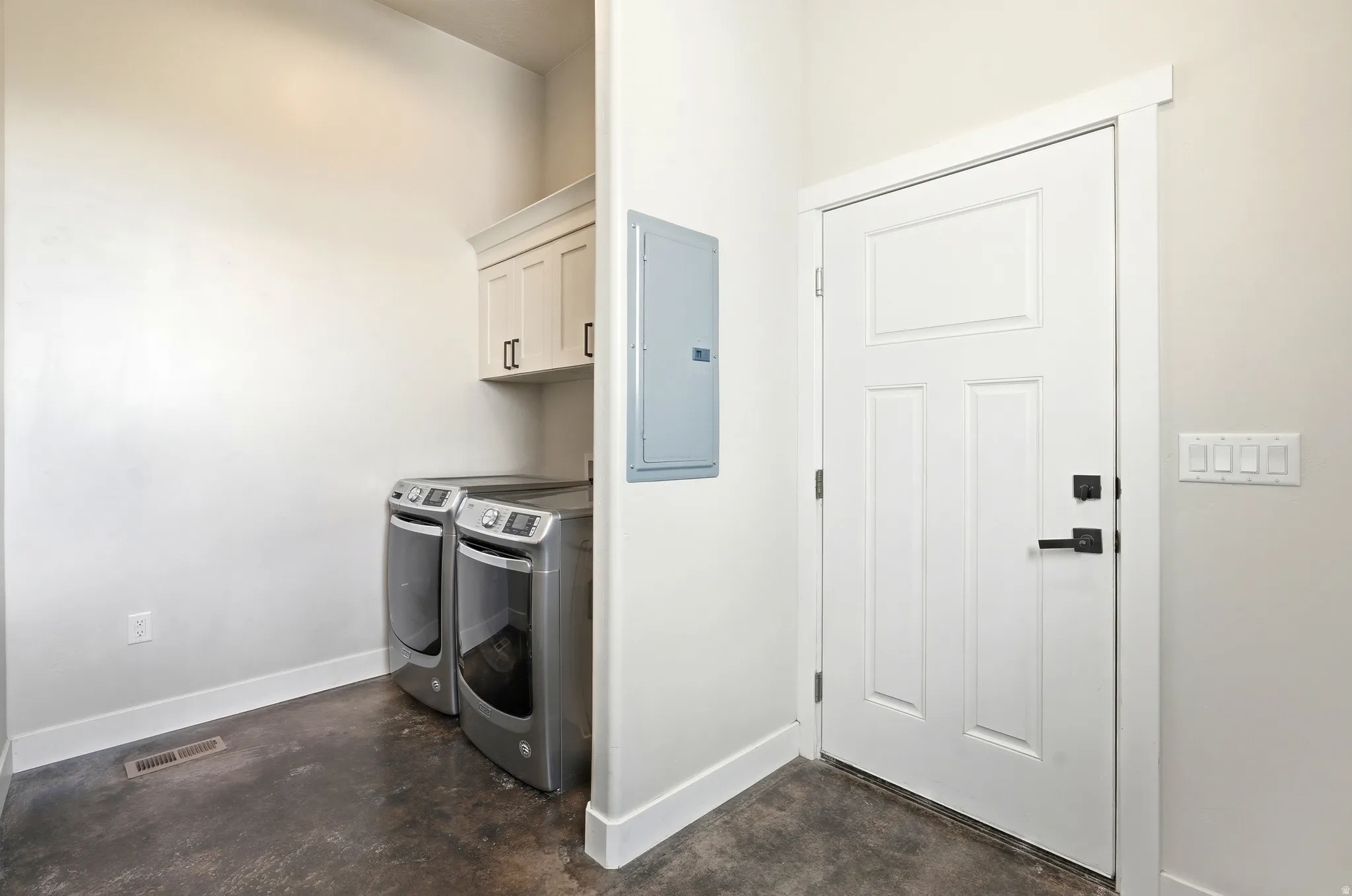 Laundry area featuring overhead cabinetry with black hardware, a panel door with black hardware, and concrete flooring. Located right off of the garage.