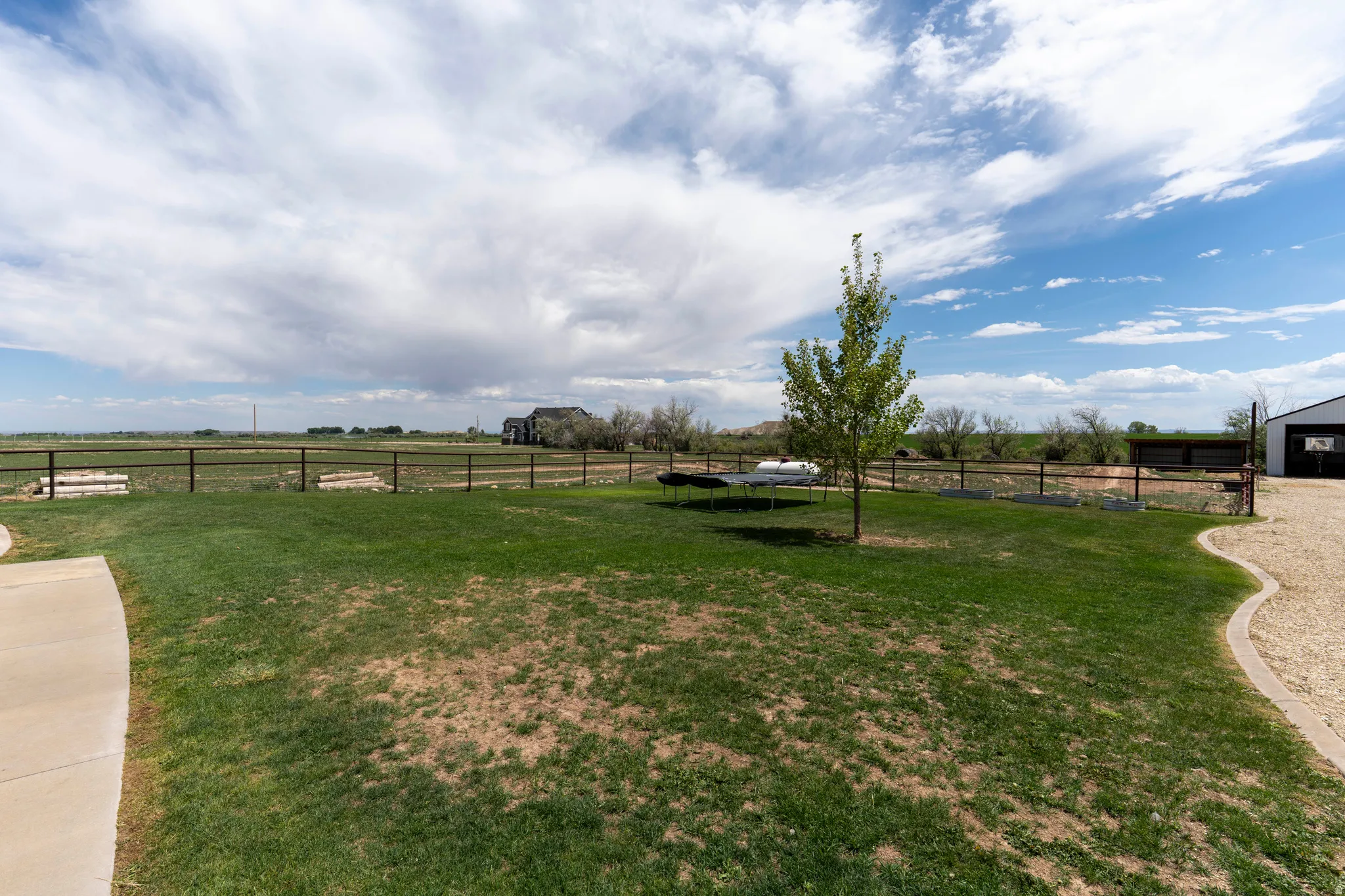 Expansive exterior featuring a manicured lawn, a young deciduous tree, a paved walkway, a gravel patio, and a perimeter fence