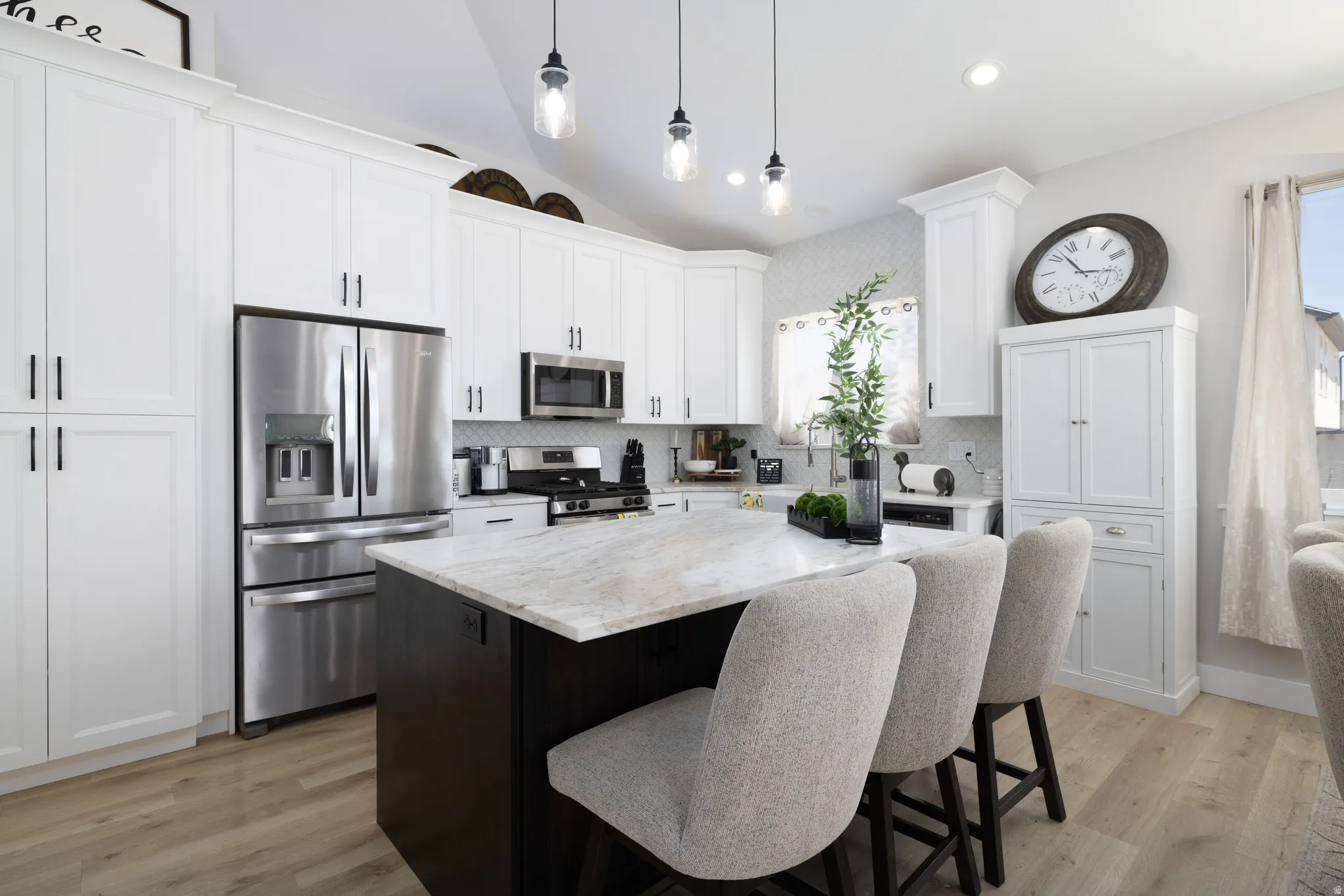 Kitchen featuring a central island with a light-toned countertop, white cabinetry with black hardware, stainless steel appliances, a white tile backsplash, and light wood-finish flooring