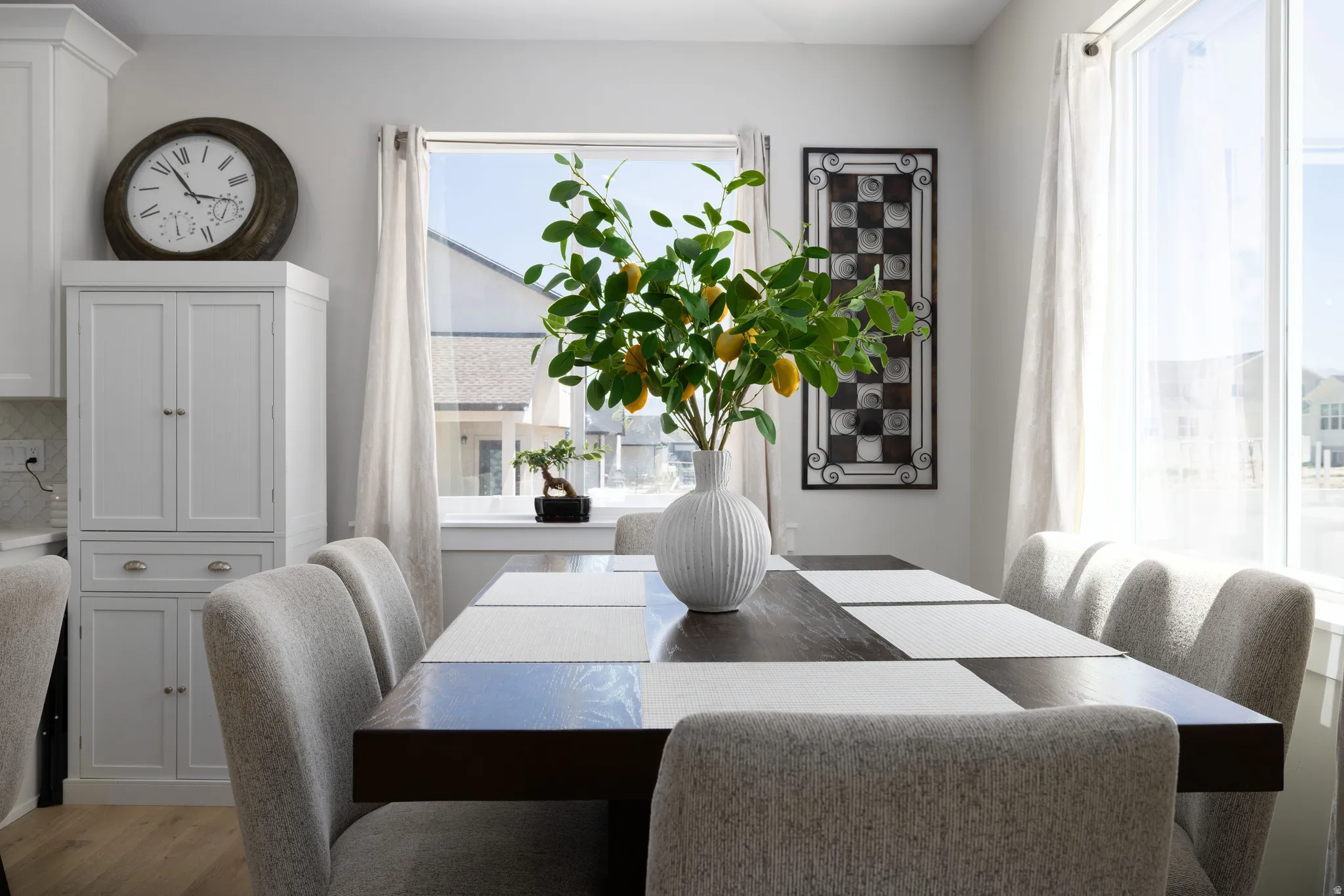 Dining area featuring a dark wood-finish table, light gray upholstered chairs, and a large window with white curtains