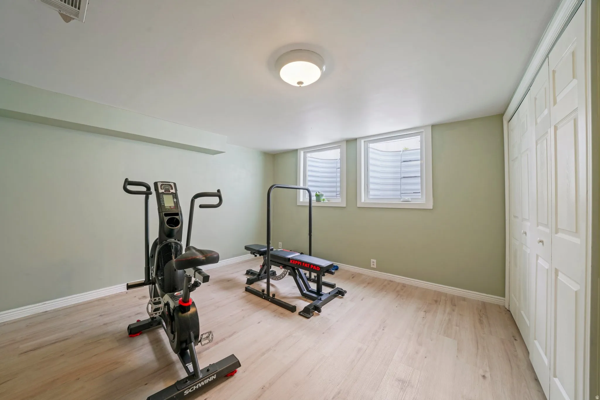 Spacious room featuring light wood-finish flooring, two egress windows with white trim, a flush-mount ceiling light fixture, and bifold closet doors