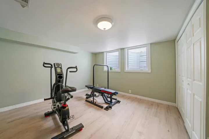 Spacious room featuring light wood-finish flooring, two egress windows with white trim, a flush-mount ceiling light fixture, and bifold closet doors