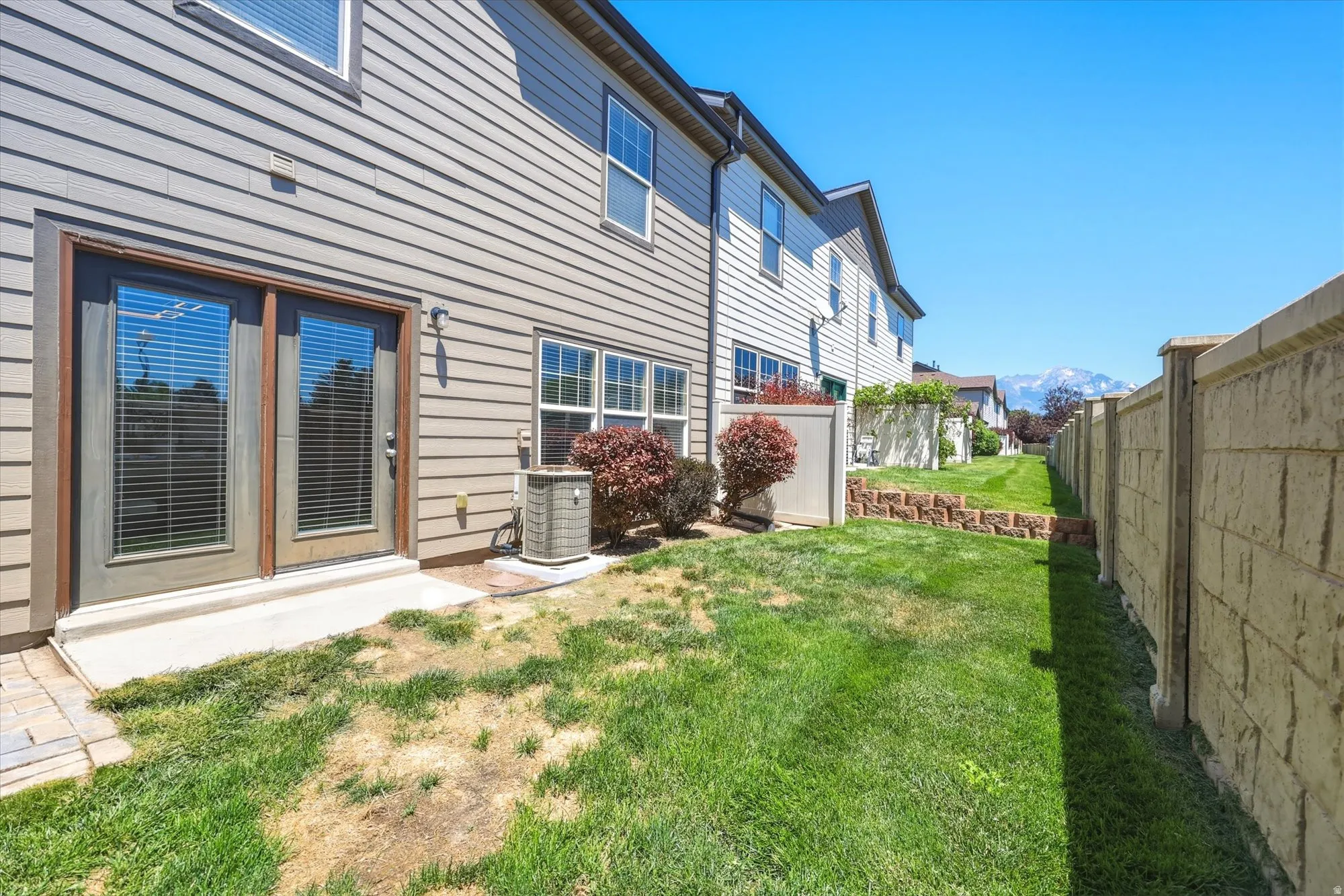 Rear exterior featuring double glass panel doors with integrated blinds, a concrete patio, and a stone block retaining wall