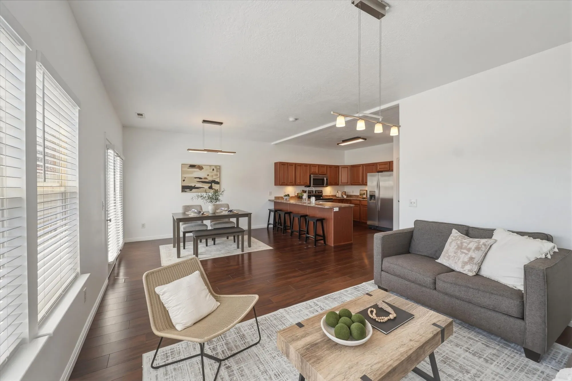 Open-concept living area featuring wood-finish flooring, a kitchen island with seating, pendant lighting, and multiple windows with blinds