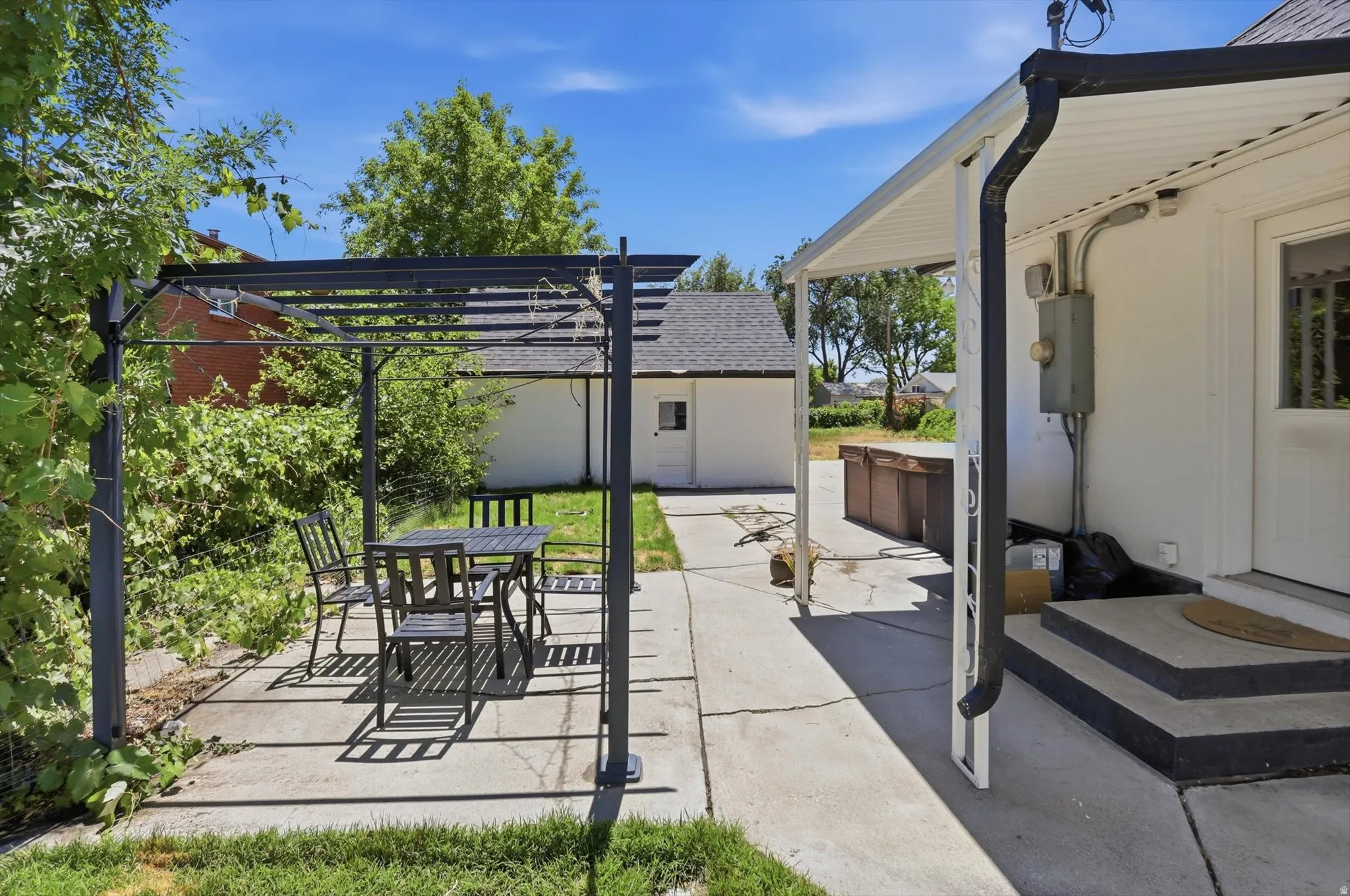 Expansive concrete patio area featuring a black metal pergola, a covered porch with white siding, a detached structure with a dark shingle roof, mature green foliage, and a separate raised wooden structure