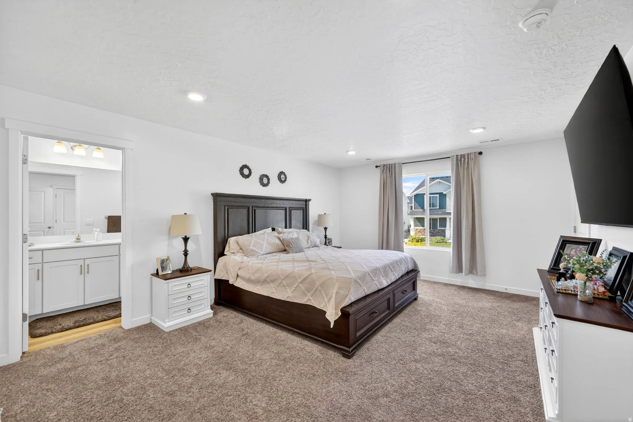 Spacious bedroom featuring plush carpeting, white walls, and a large window with neutral drapes